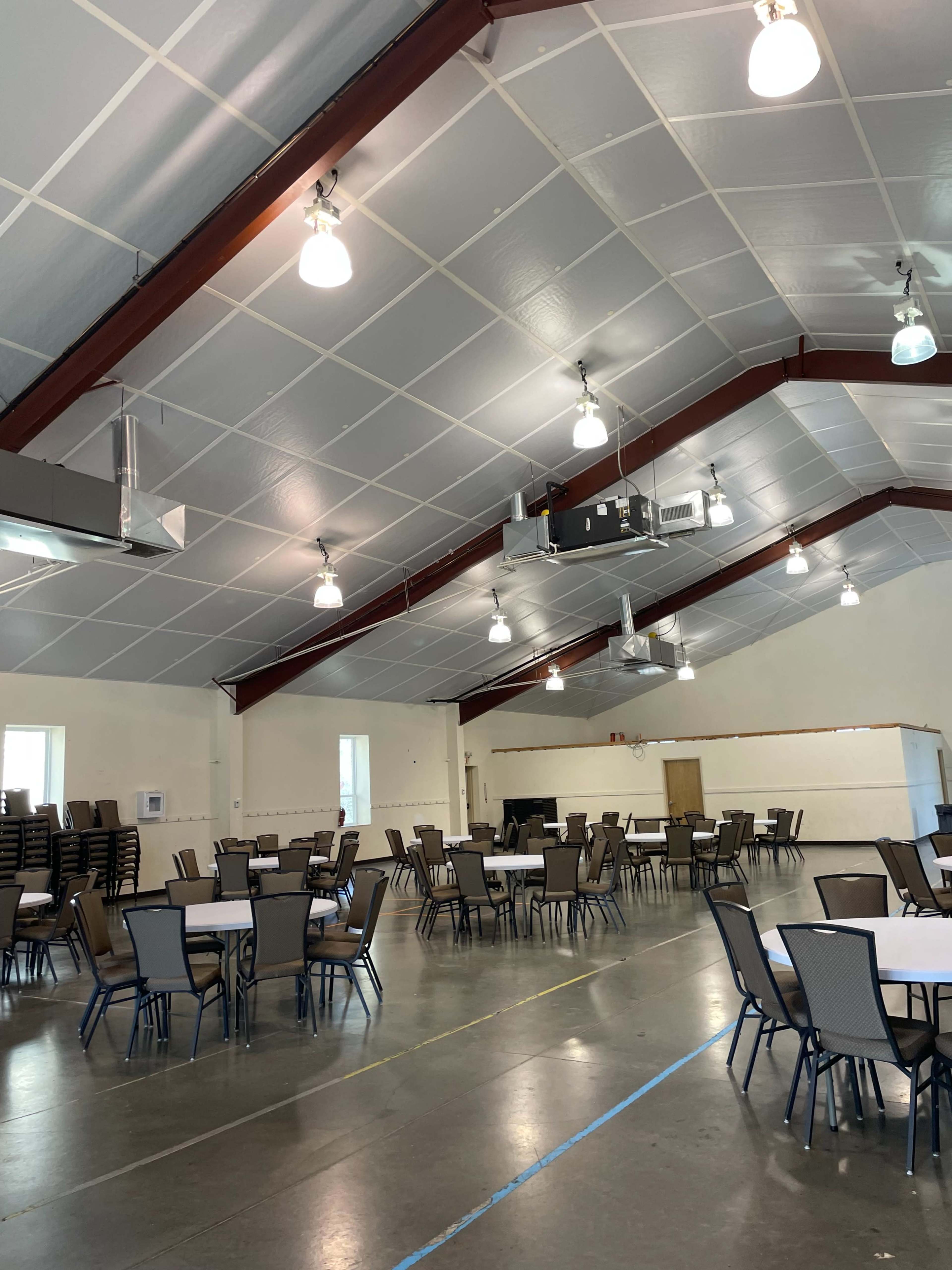 The image shows a large indoor event space with several round tables and chairs arranged neatly on a smooth floor beneath a high ceiling with overhead lights.