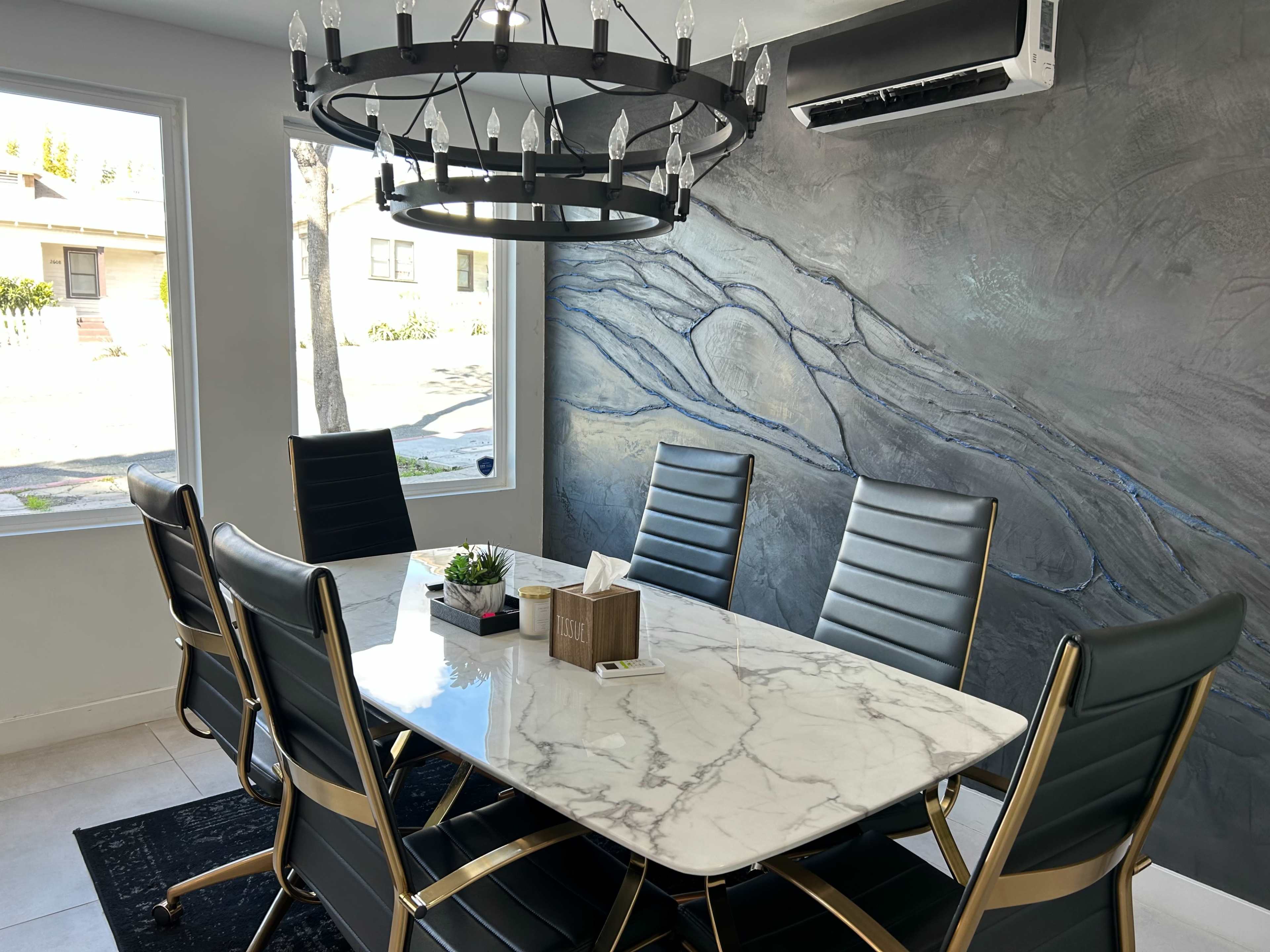 A modern dining room features a marble table surrounded by black leather chairs, with a large chandelier above and a textured wall mural.