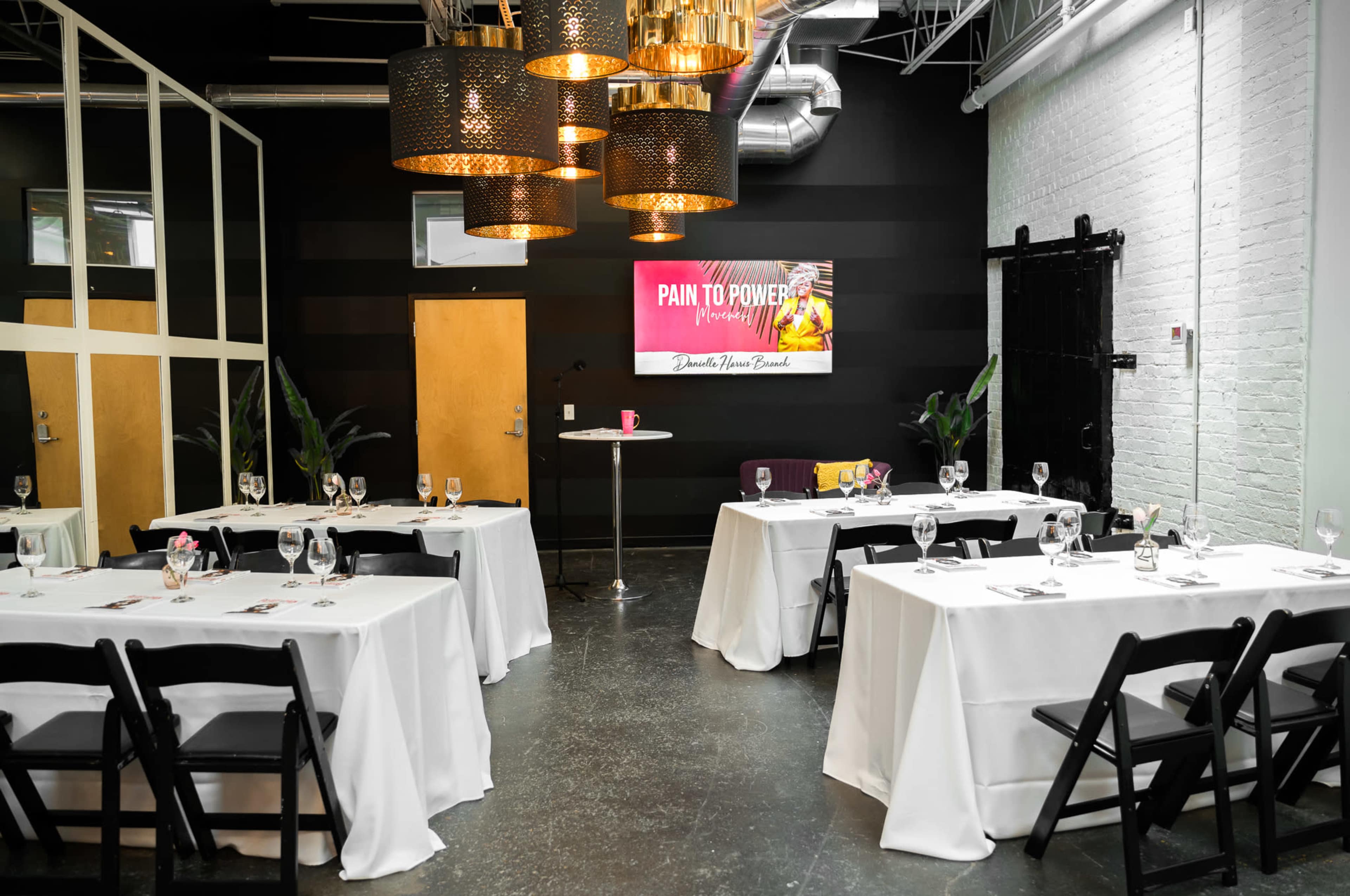 A stylish event space with black walls, white tables set with tableware, and pendant lighting, featuring a screen displaying "Pain to Power."