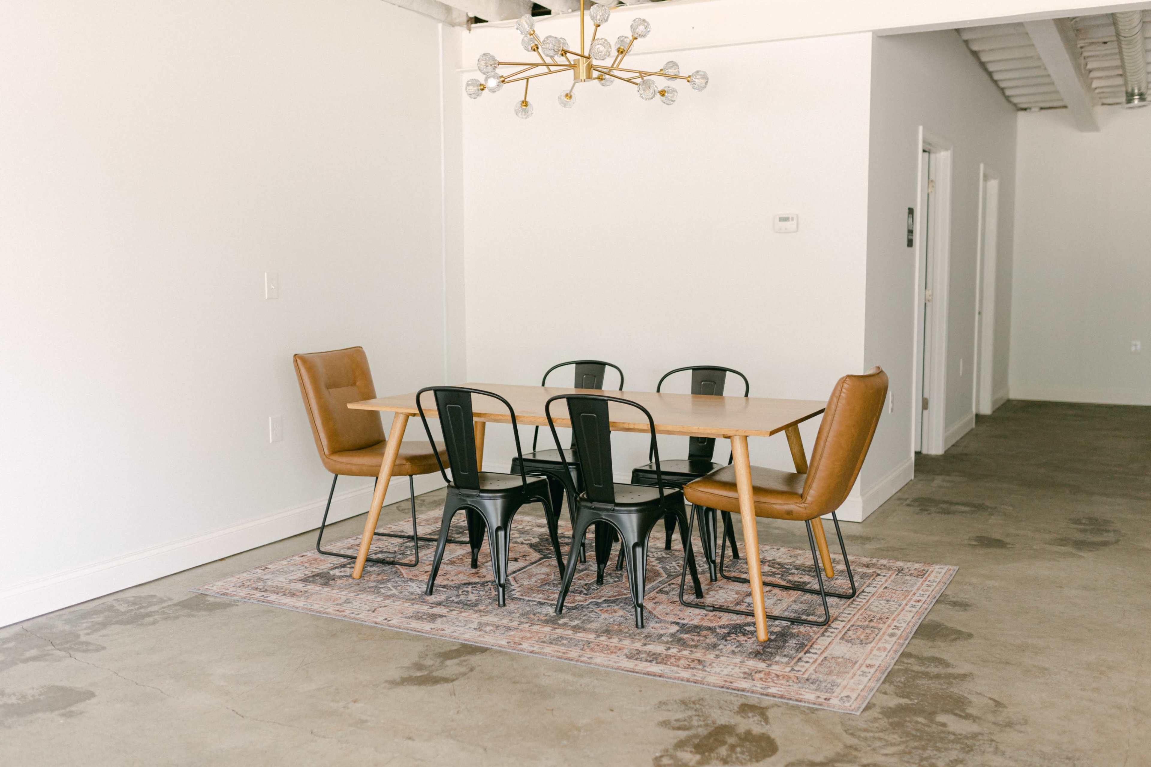 A wooden dining table surrounded by black metal and brown leather chairs sits on a patterned rug in a minimalist room with concrete flooring.