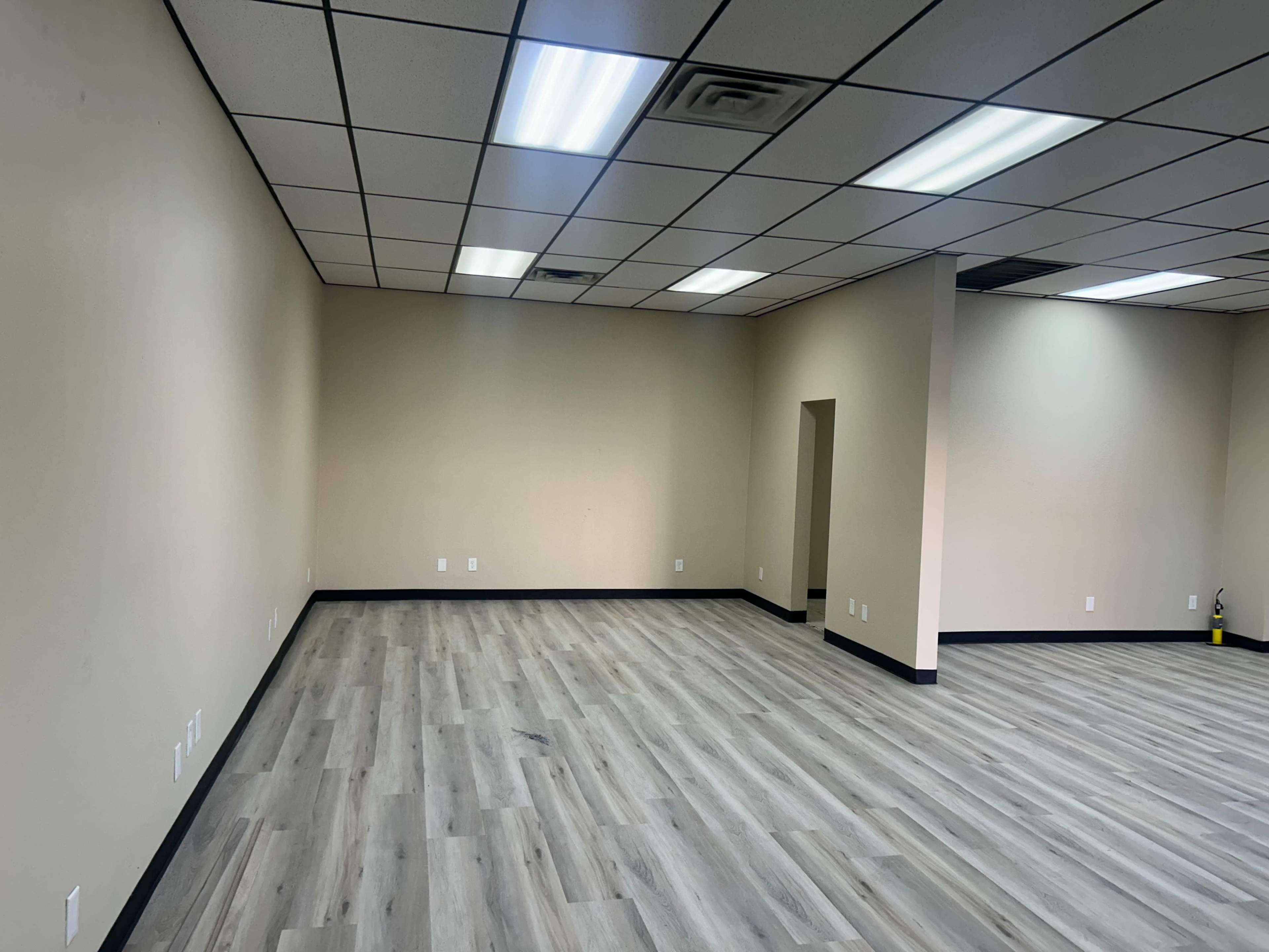 The image shows a vacant room with light-colored walls, a hardwood floor, and fluorescent ceiling lights.