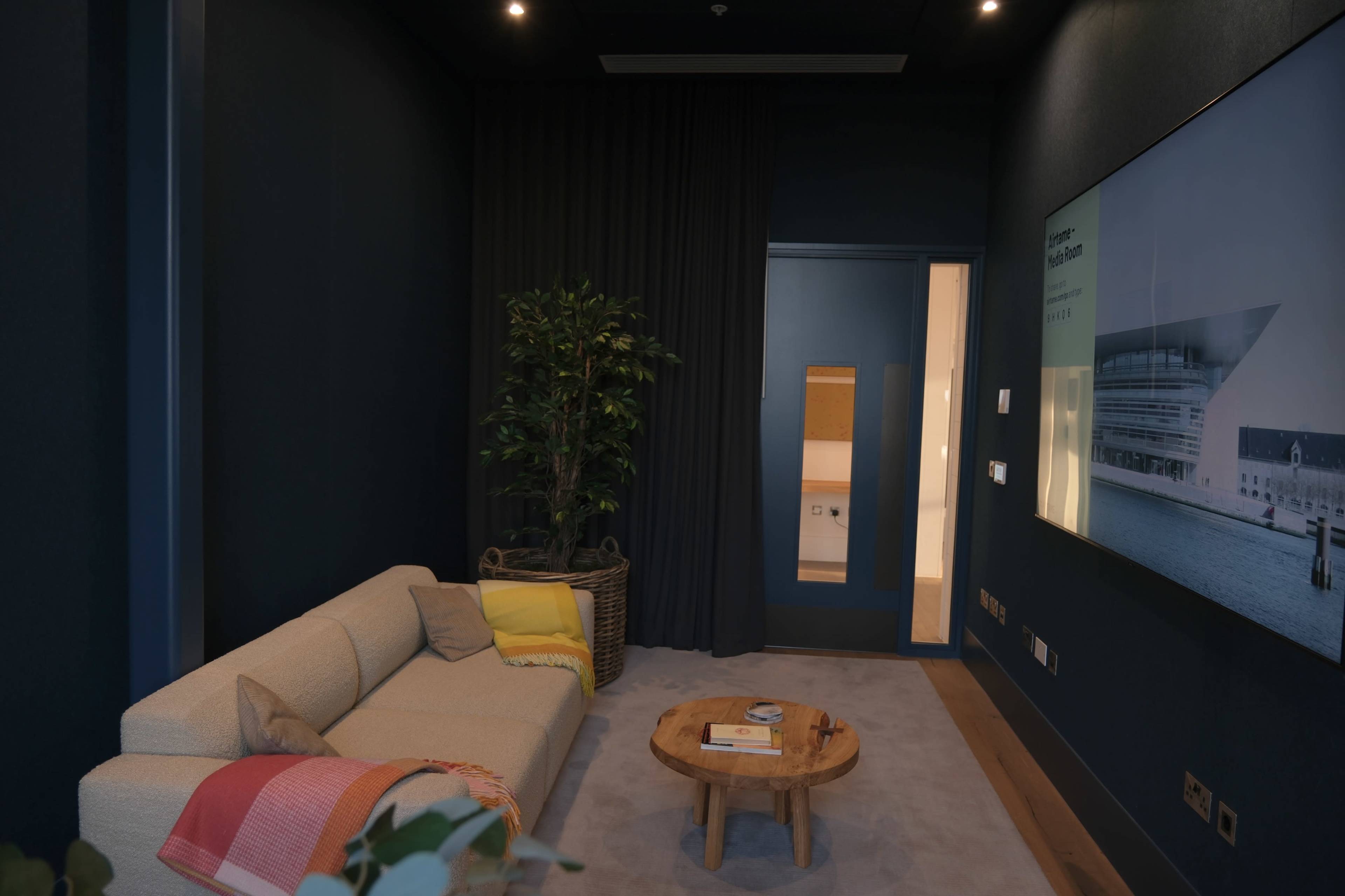 A small, modern lounge area featuring a beige sofa, a round wooden coffee table, a potted plant, and dark walls with a partially visible door in the background.