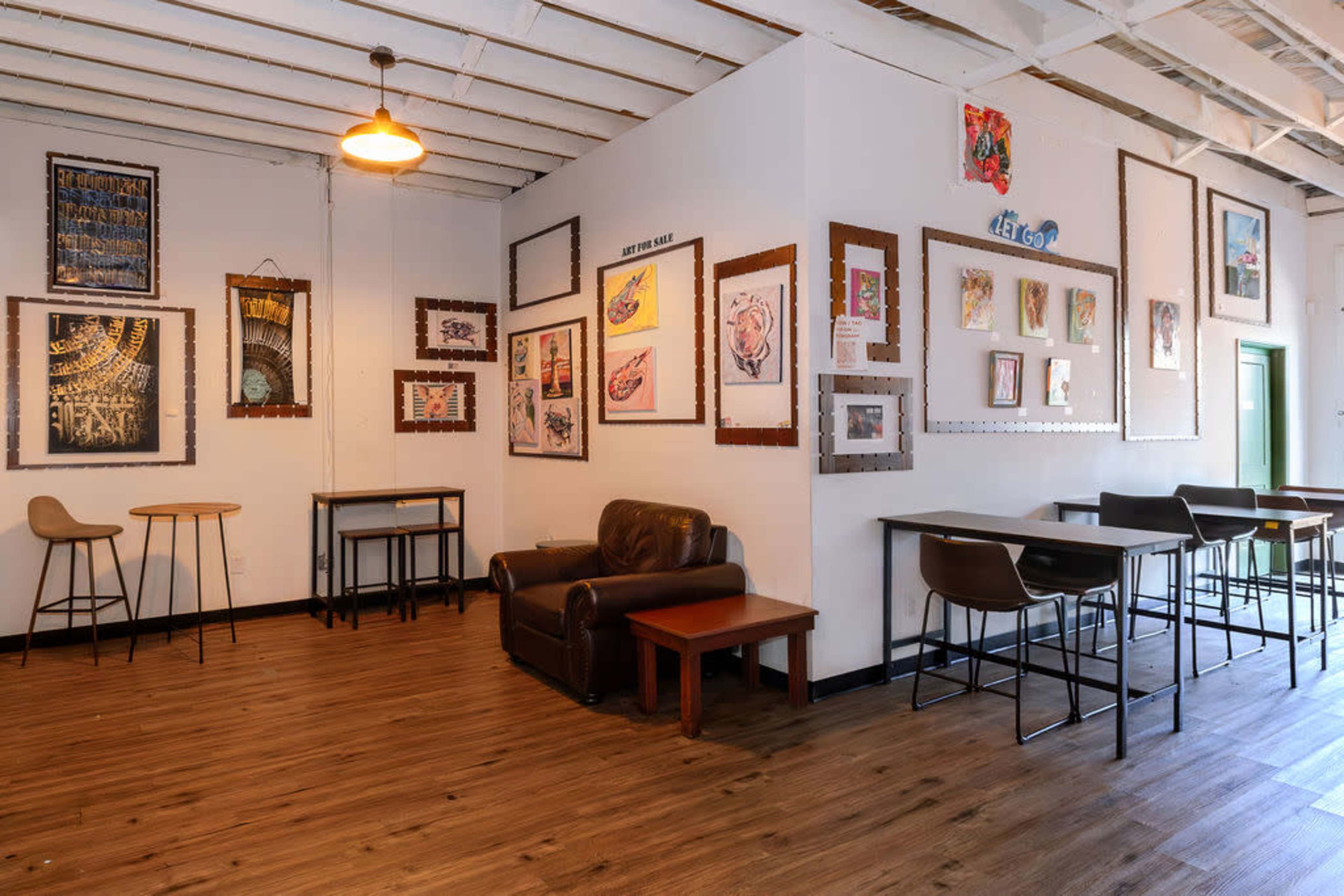 The image shows an inviting café interior with wooden floors, a mix of table seating, a leather couch, and various framed artworks displayed on the walls.