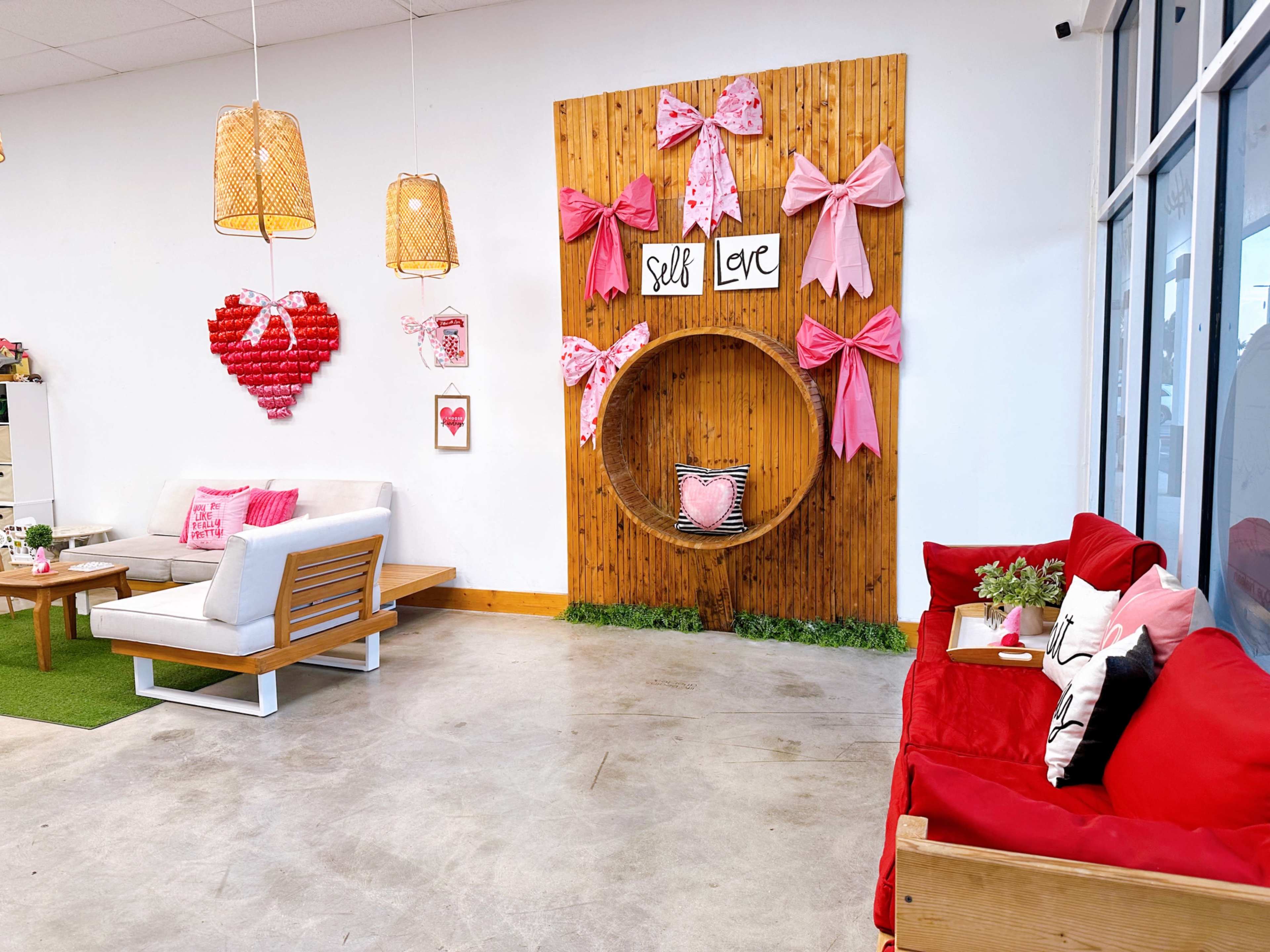 The image shows a decorated interior space with a wooden accent wall featuring a circular display, pink bows, and heart-shaped decor, along with seating areas arranged with light and dark-colored couches.