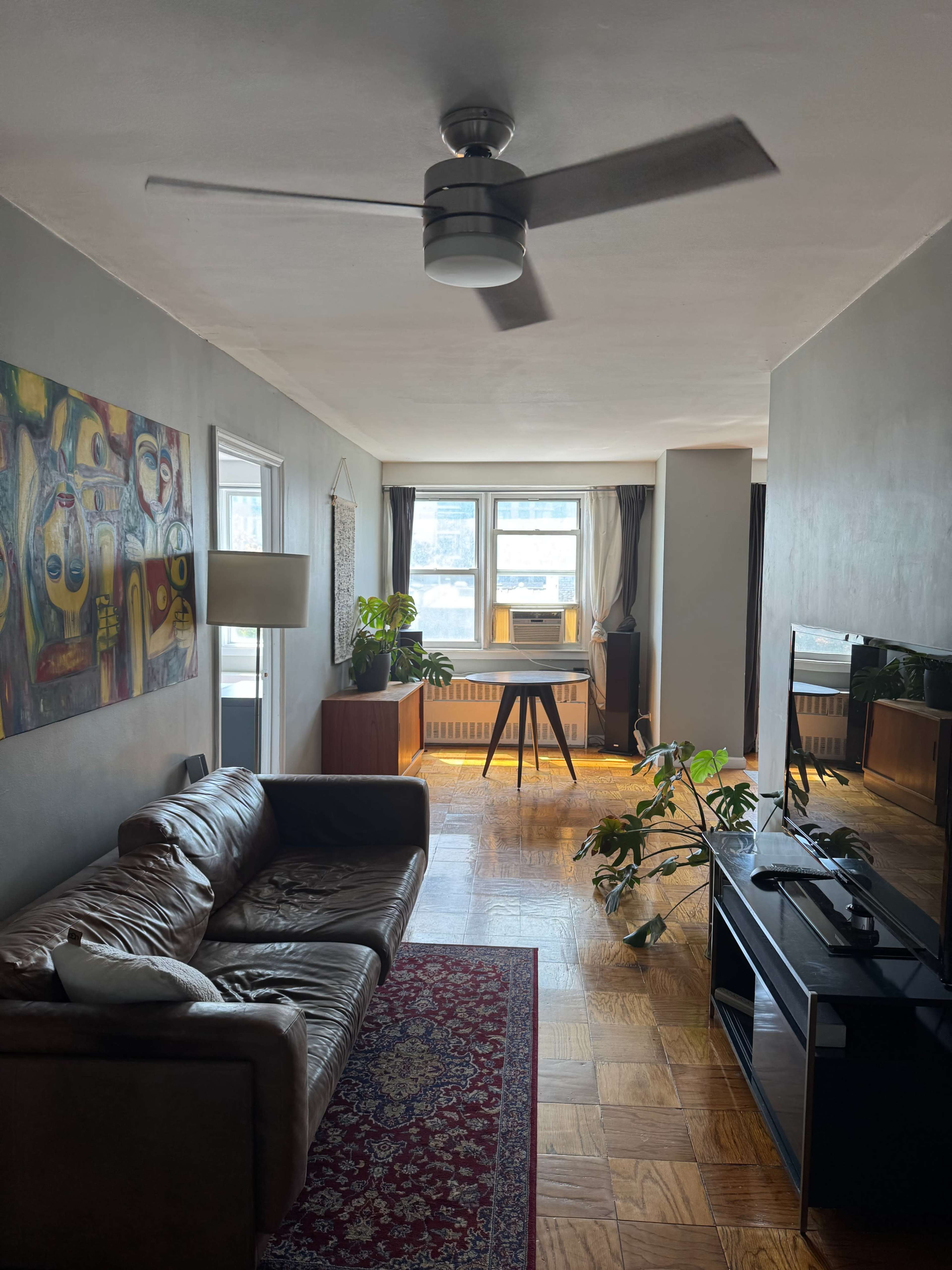 The image shows a living room featuring a leather sofa, a coffee table, a large window with natural light, and a television stand with a plant nearby.