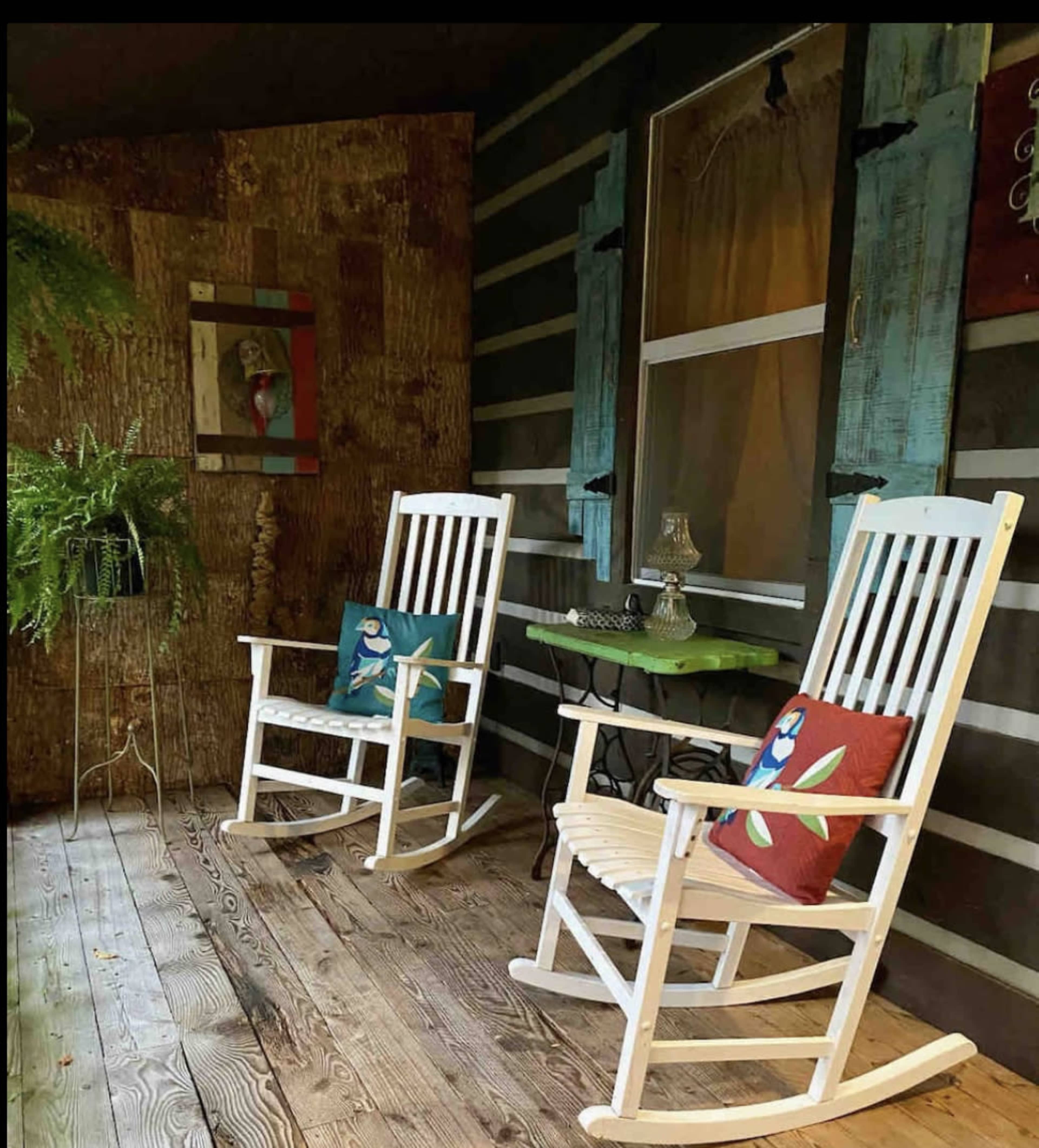 Two white rocking chairs with decorative cushions placed on a wooden porch beside a small green table and a fern.