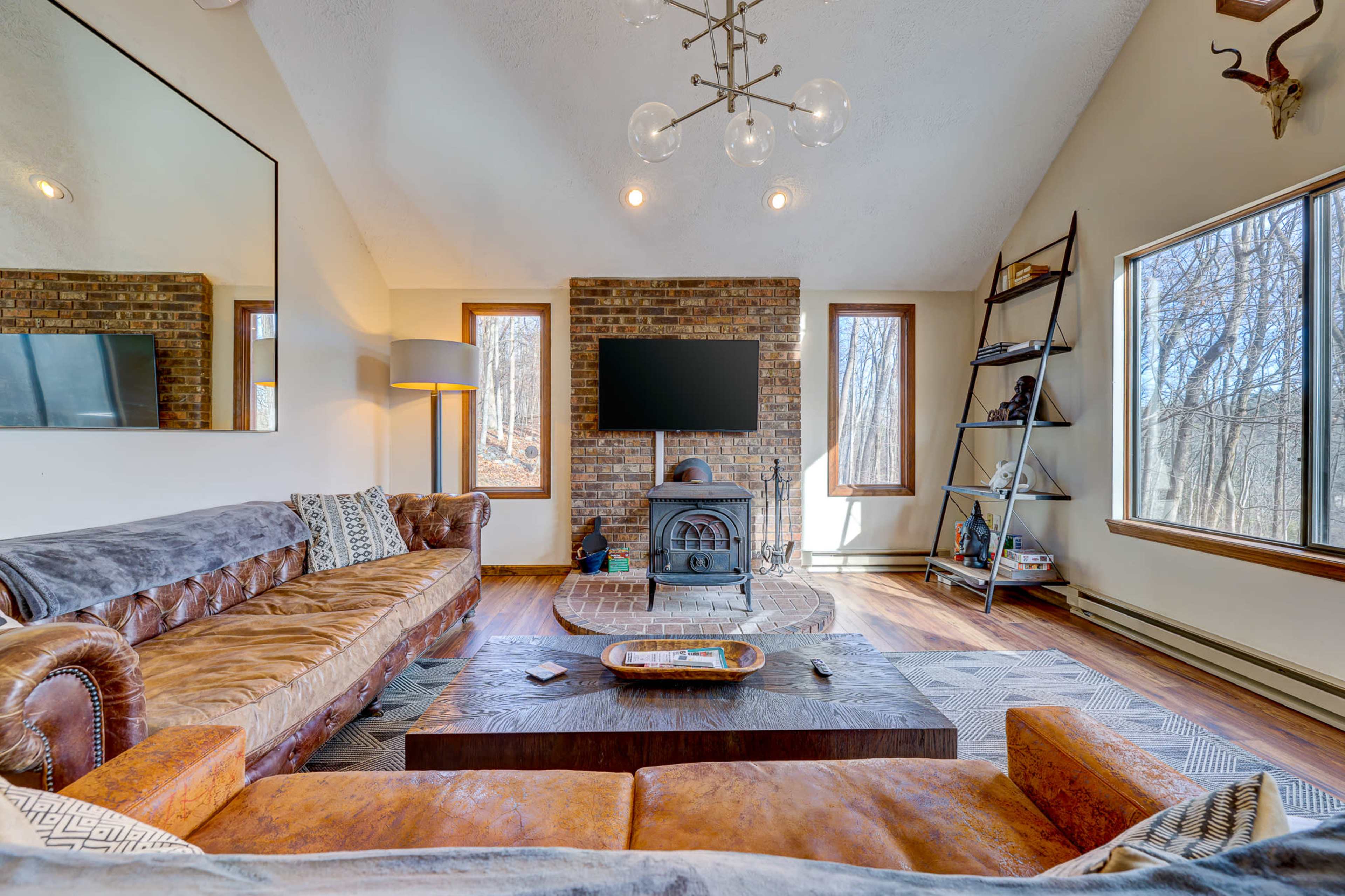 The image shows a modern living room featuring a leather sofa, a wooden coffee table, a wood-burning stove, and large windows overlooking a wooded area.