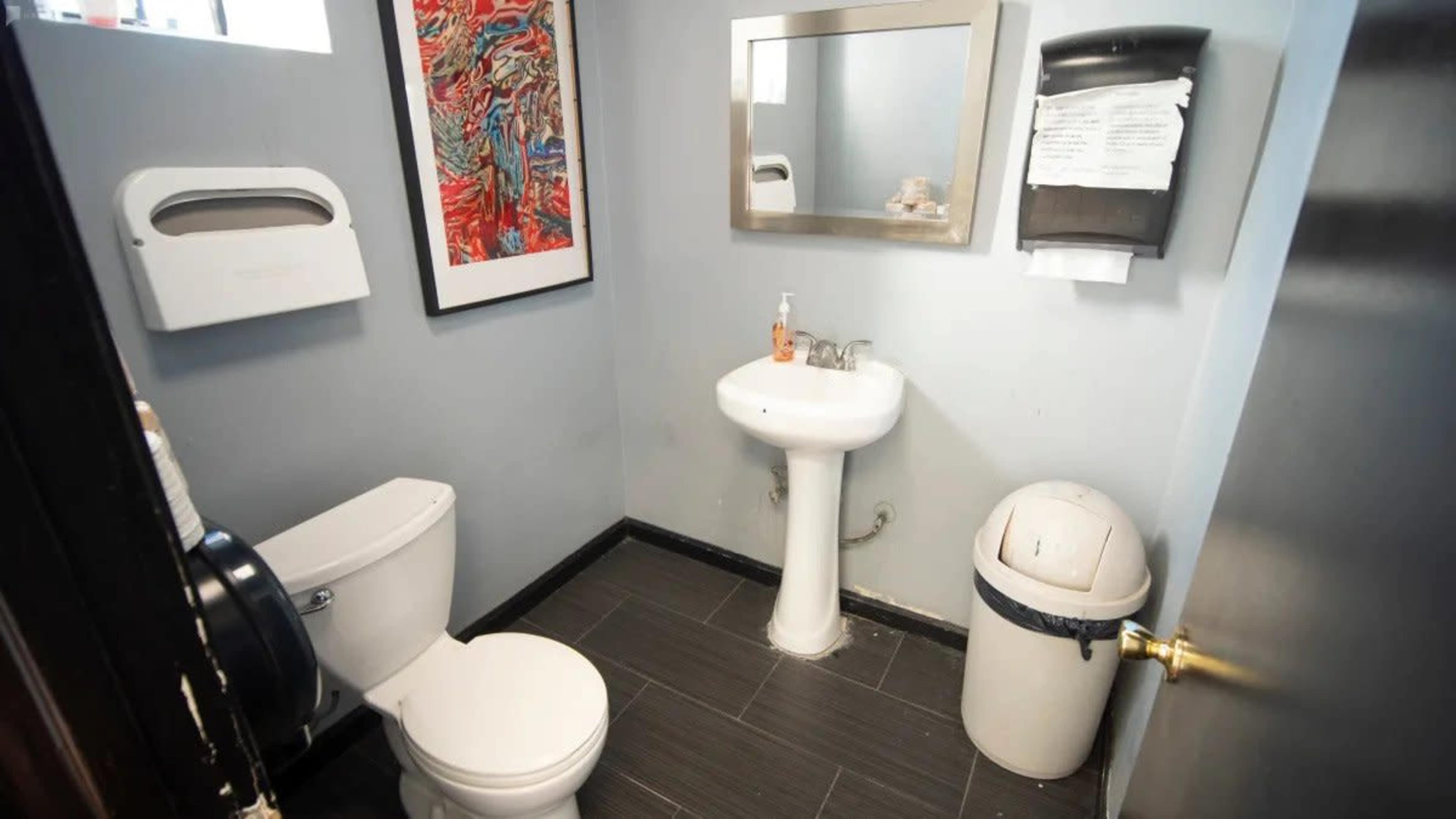 A small bathroom featuring a toilet, a sink, a mirror, a wall-mounted soap dispenser, and a trash bin.