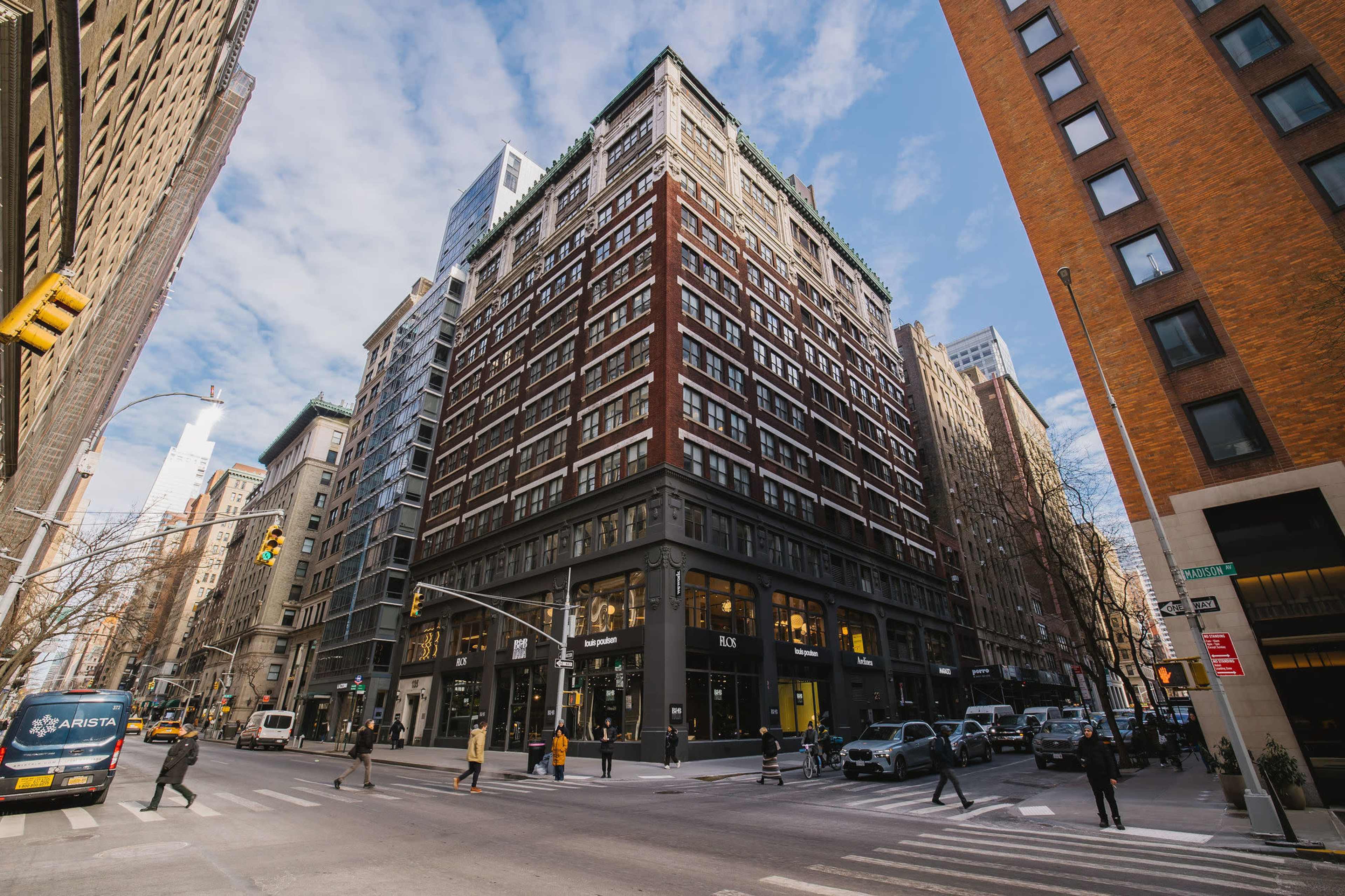 A busy urban intersection features a tall brick building on the corner, with pedestrians crossing and vehicles parked along the street.