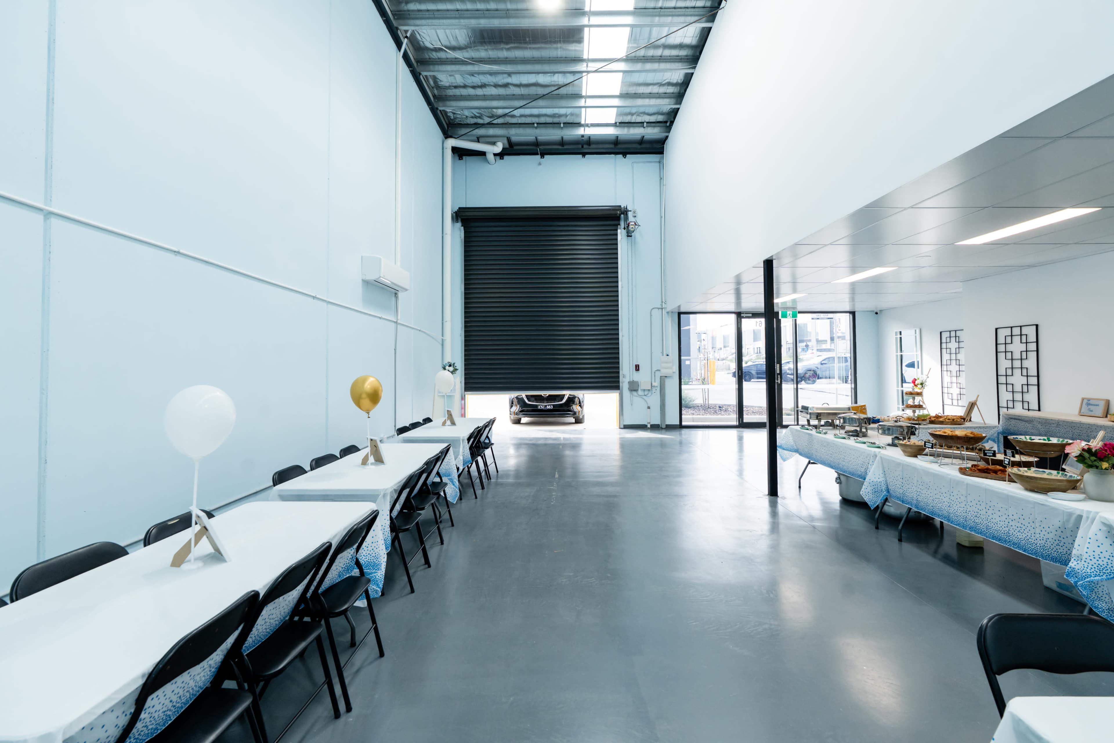 The image shows a spacious indoor event area with several long tables set up, a large roll-up door open at the far end, and food laid out on the tables.