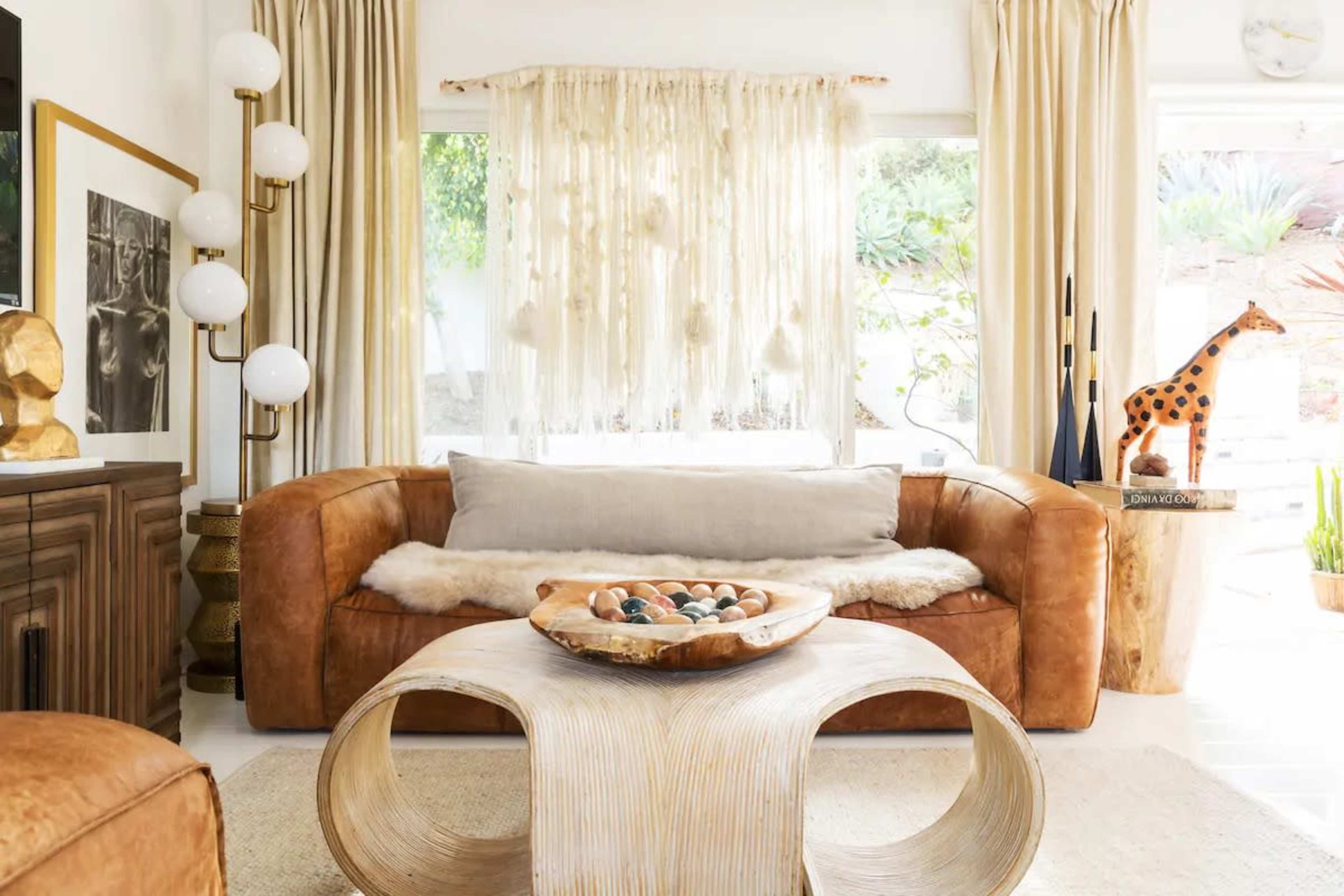 The living room features a leather sofa, a round wooden coffee table, and large windows with decorative curtains, accented by a tall giraffe figure and various decorative objects.