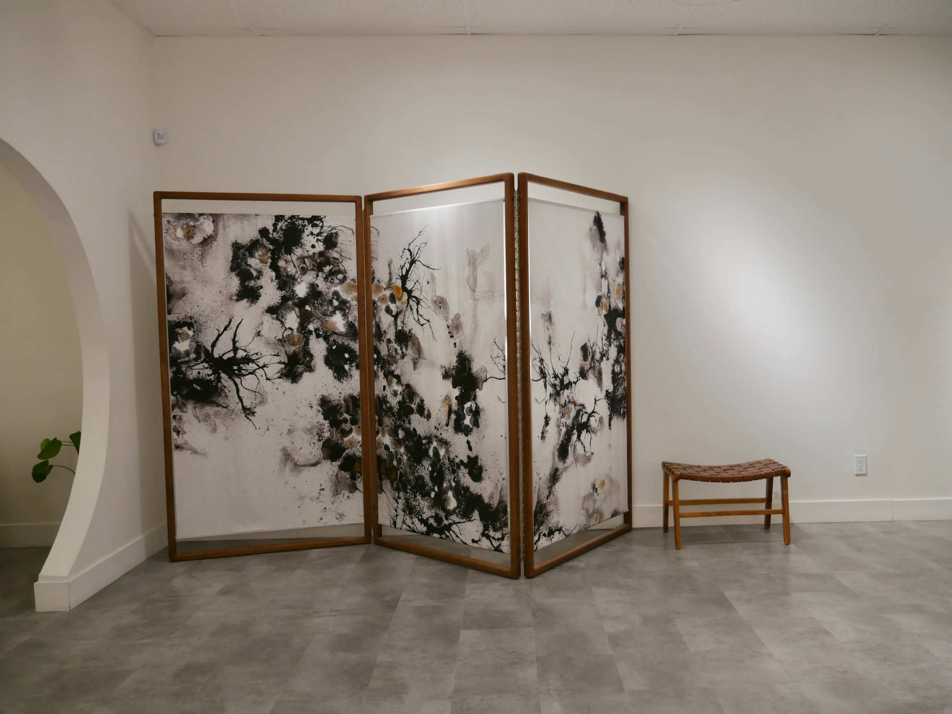 A wooden room divider with abstract artwork stands in a minimalist interior beside a small wooden bench.