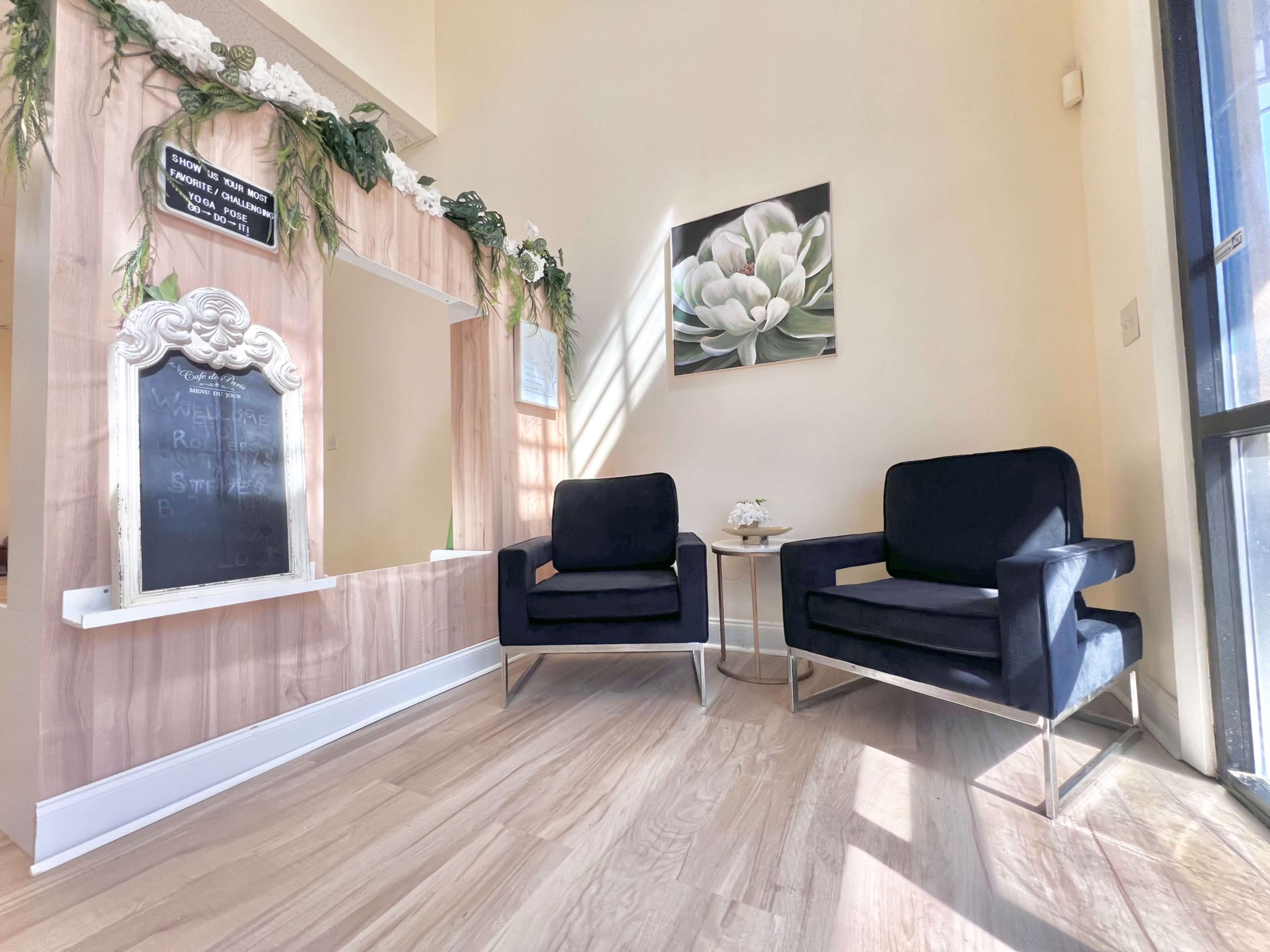 The image shows a bright, minimalist interior space featuring two black chairs facing an ornate mirror framed with greenery and a wall-mounted artwork of a flower.