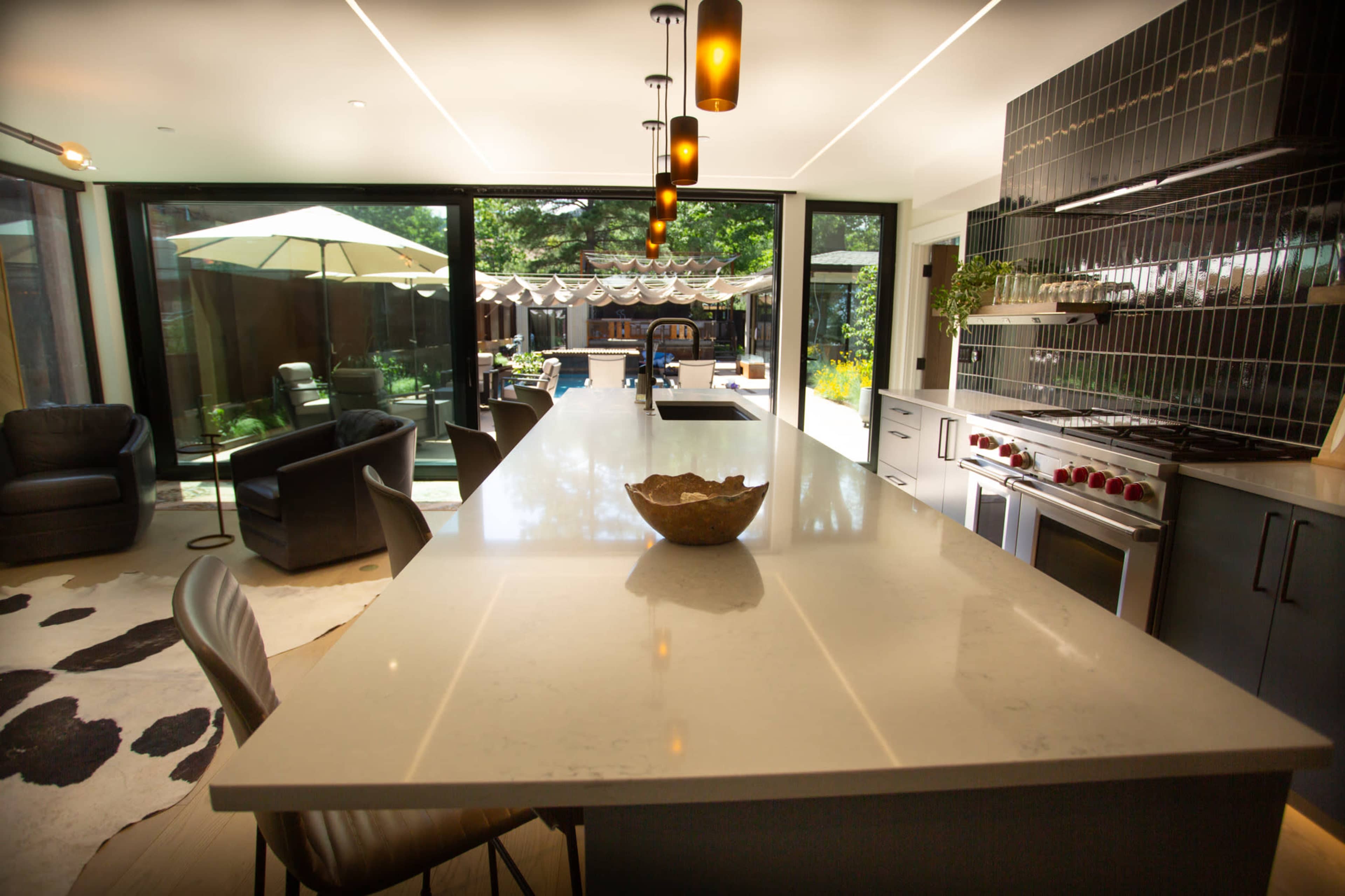 The image shows a modern kitchen with a large island, black tile backsplash, and an adjacent dining area visible through glass walls, featuring outdoor seating under an umbrella.