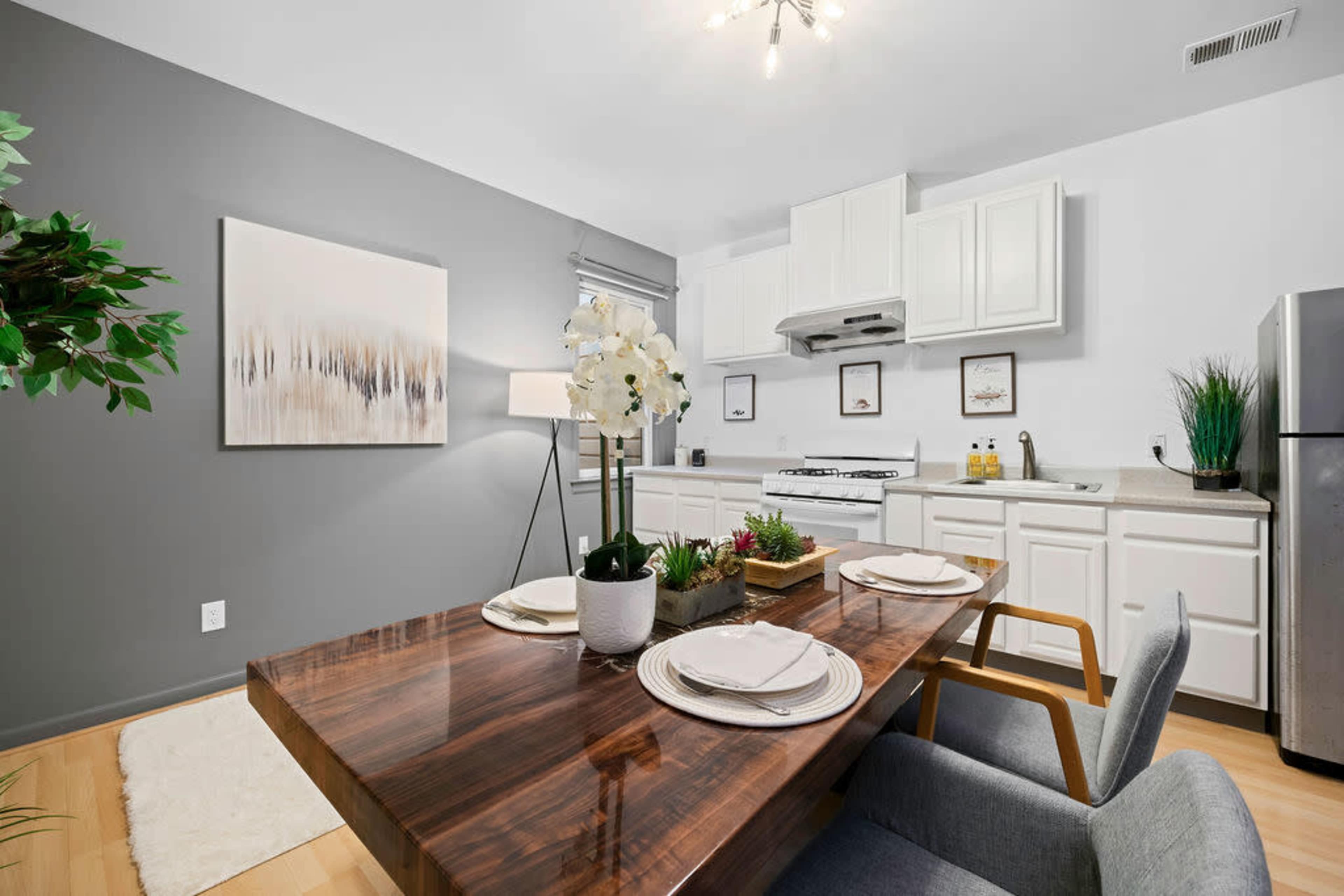 A modern kitchen and dining area features a wooden table set for four, with white cabinetry, a gas stove, and decorative plants.