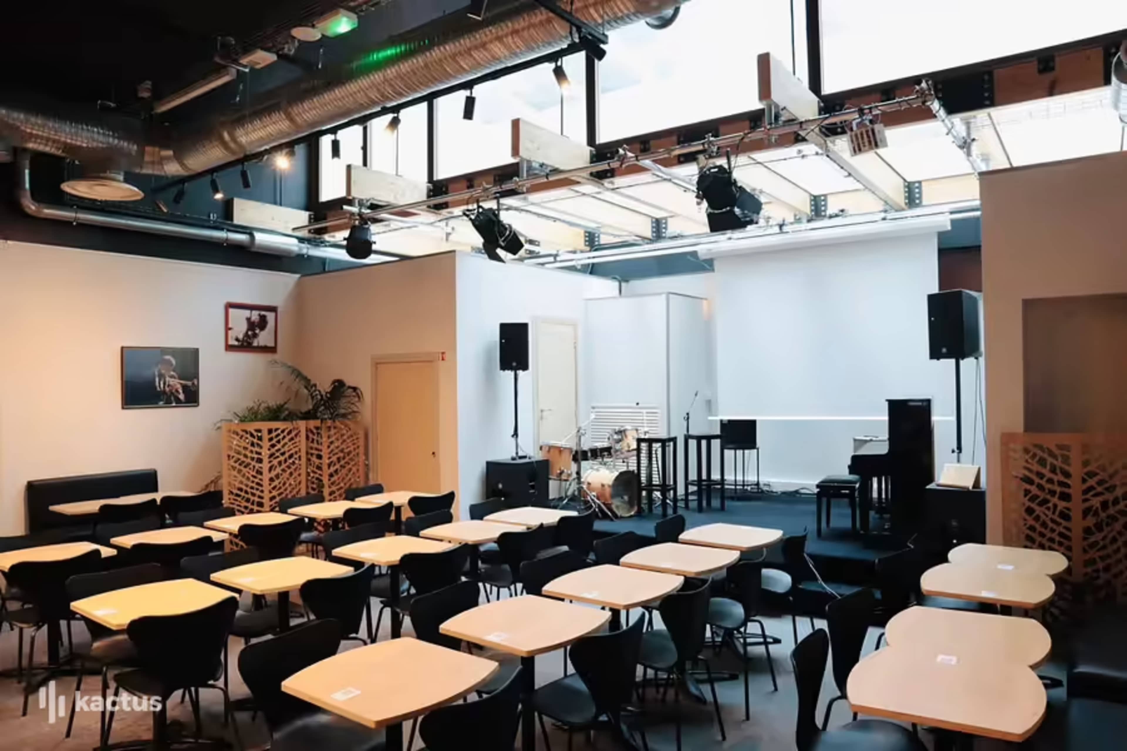 A partially set-up performance space with empty tables, a stage featuring a piano and drum kit, and sound equipment in a modern interior.