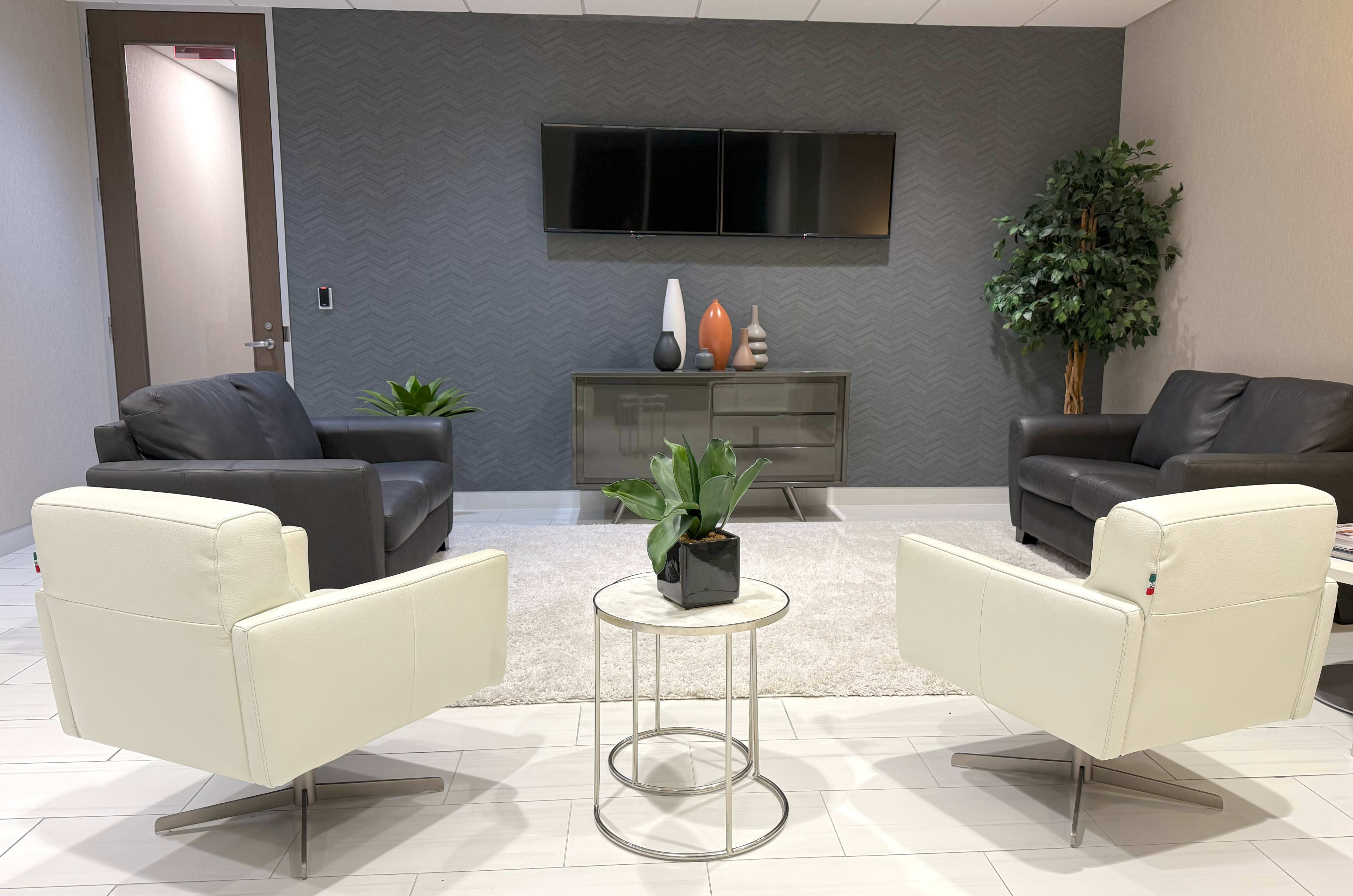 A modern waiting area features two black sofas, two white chairs, a circular coffee table with a plant, and a console table displaying decorative vases, set against a gray wall.