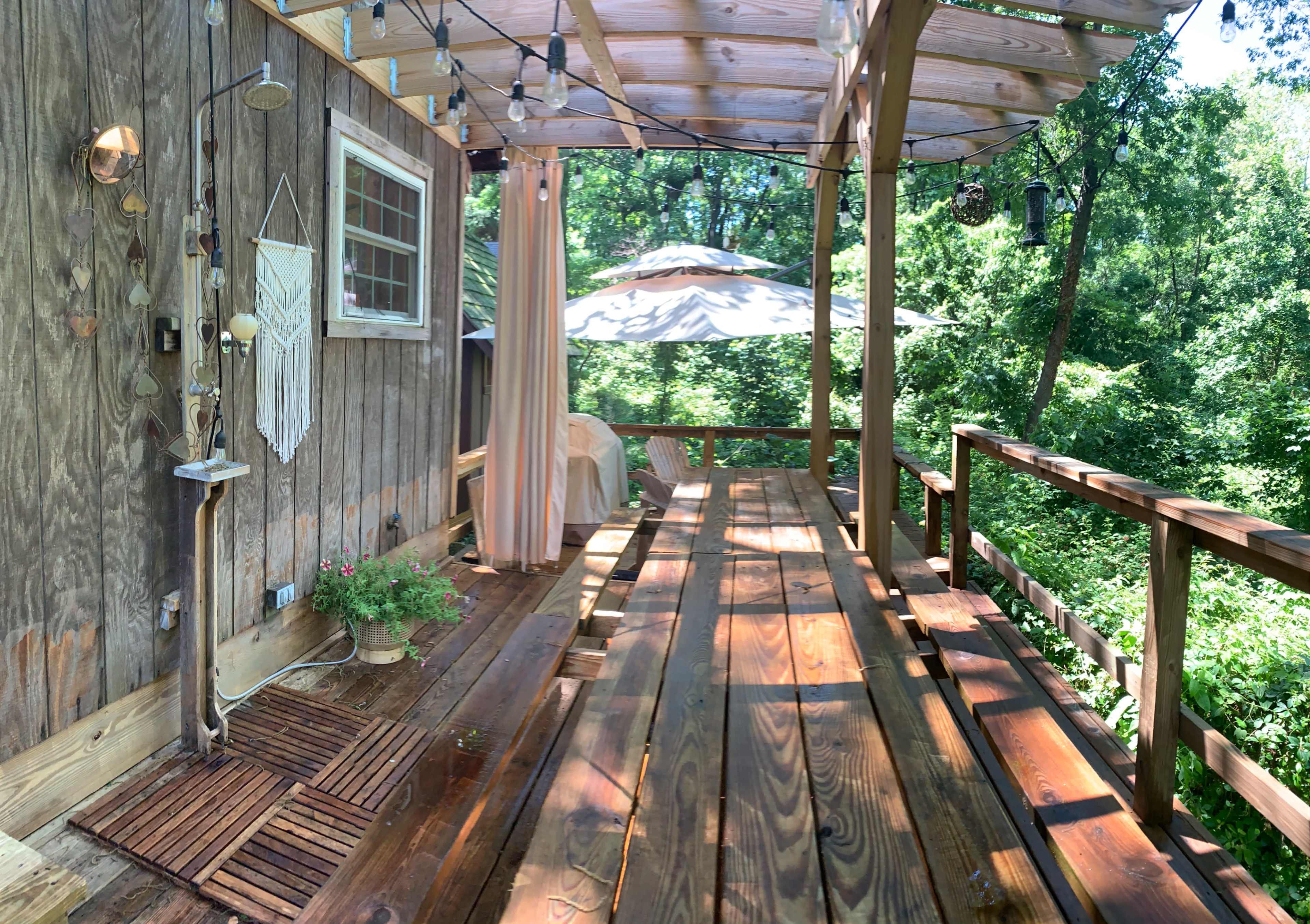 A wooden deck with a parasol, surrounded by trees and featuring string lights and a potted plant.