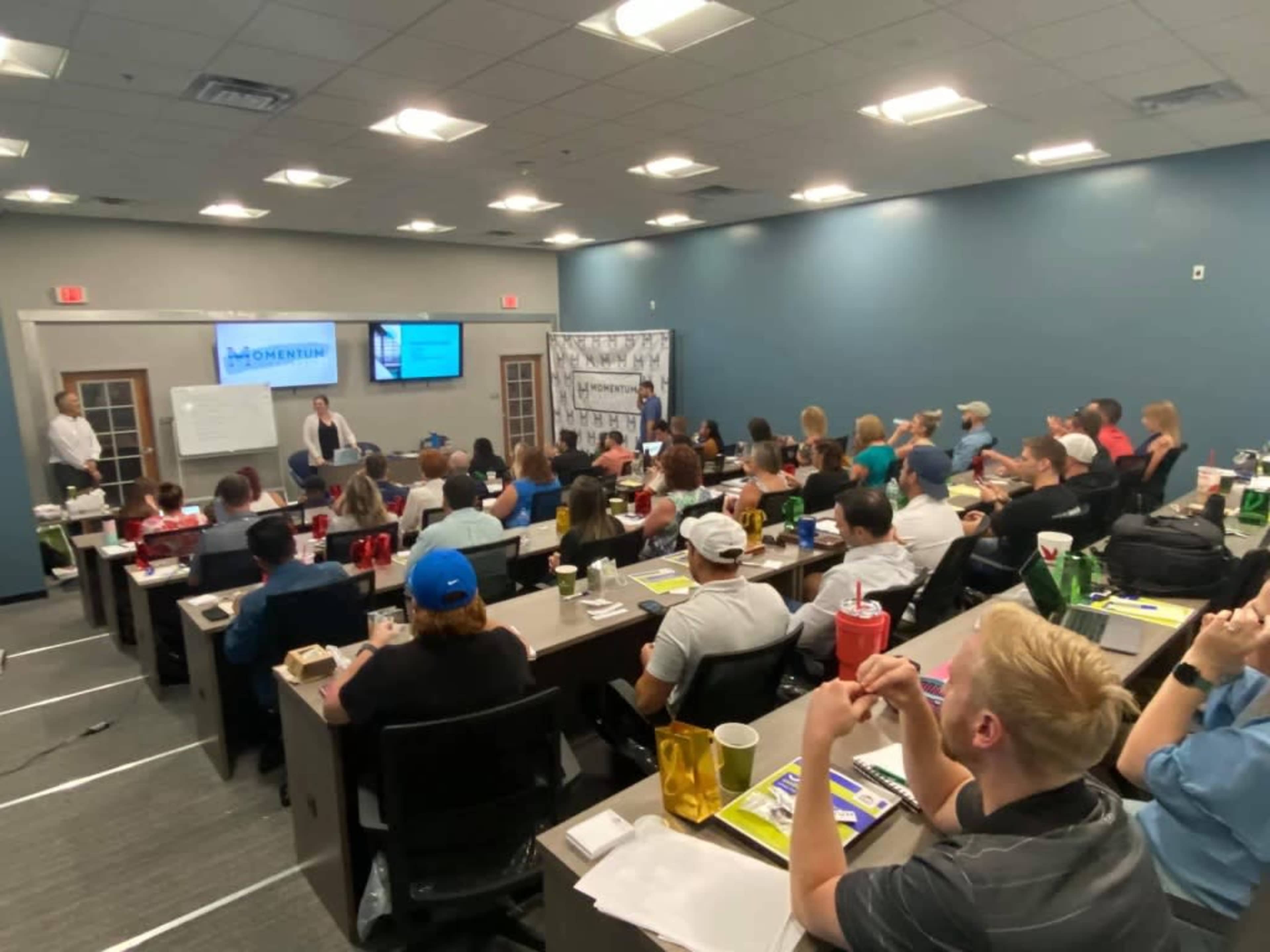 A classroom filled with attendees participates in a presentation with a speaker at the front and a projector displaying information.