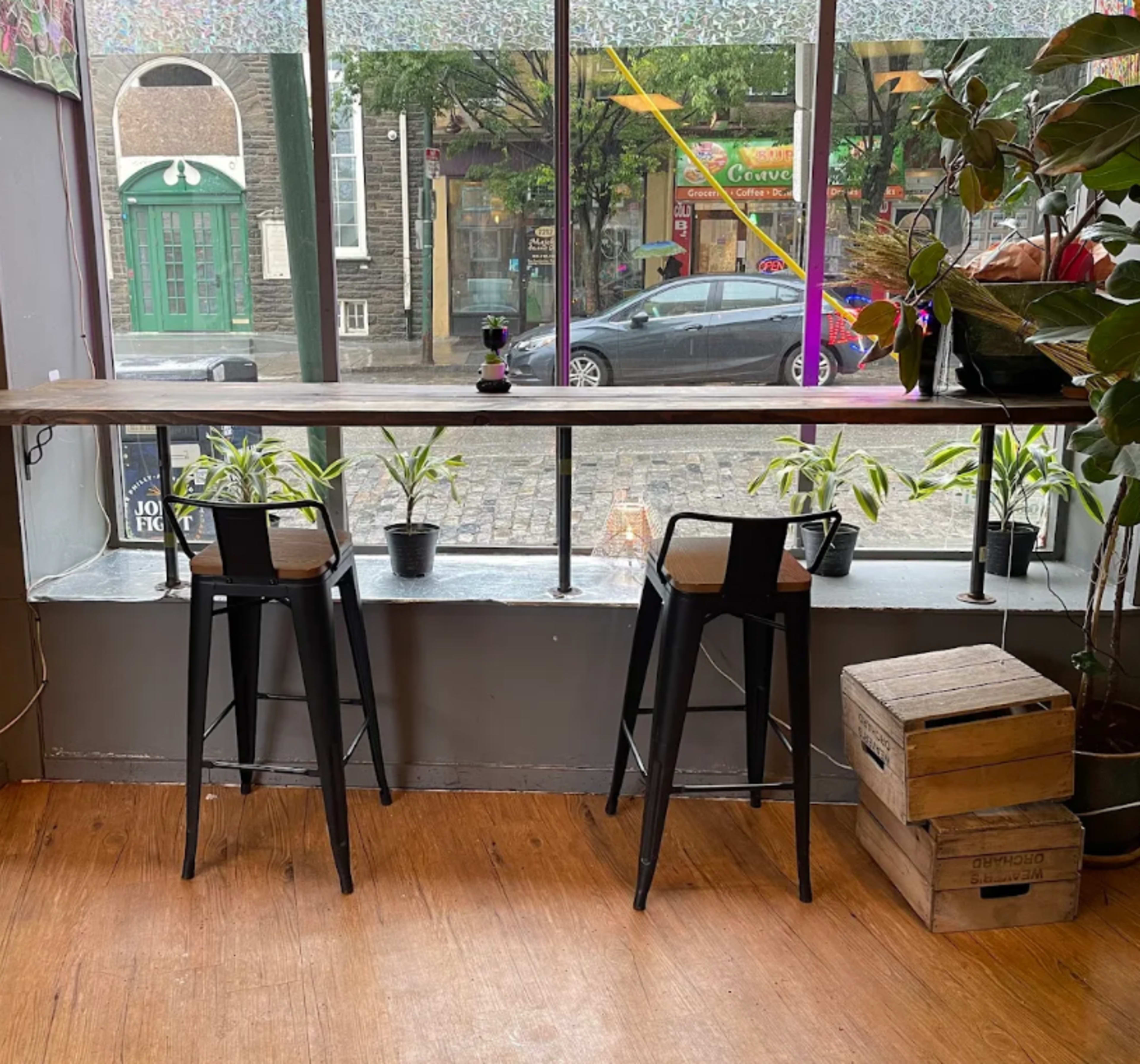 The image shows a cozy café interior with two black bar stools positioned at a window counter, alongside potted plants and a view of a street outside.