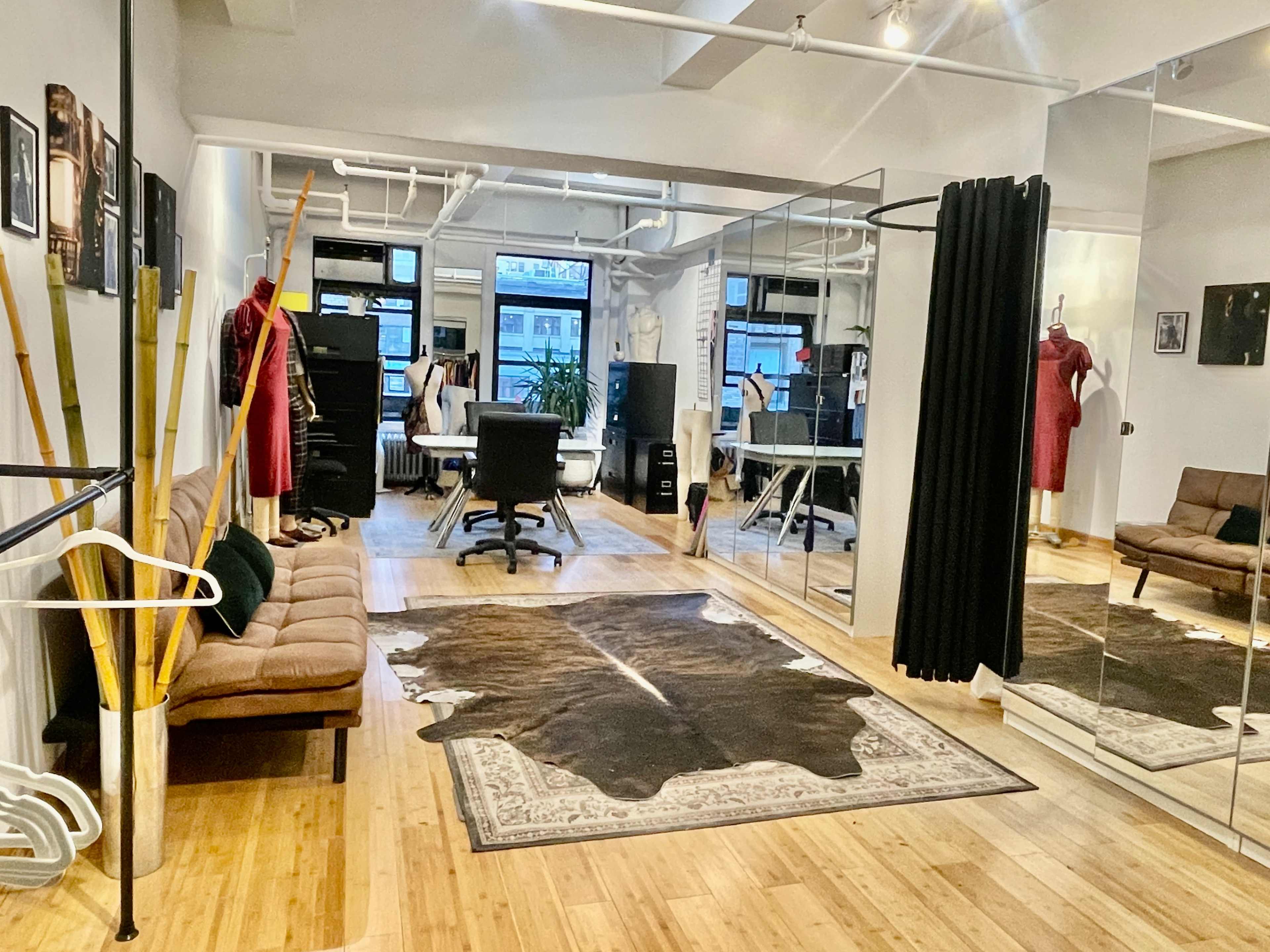 The image shows a stylish interior space featuring mirrors, a desk and chair, a sofa, and a clothing rack, with wooden flooring and a cowhide rug.