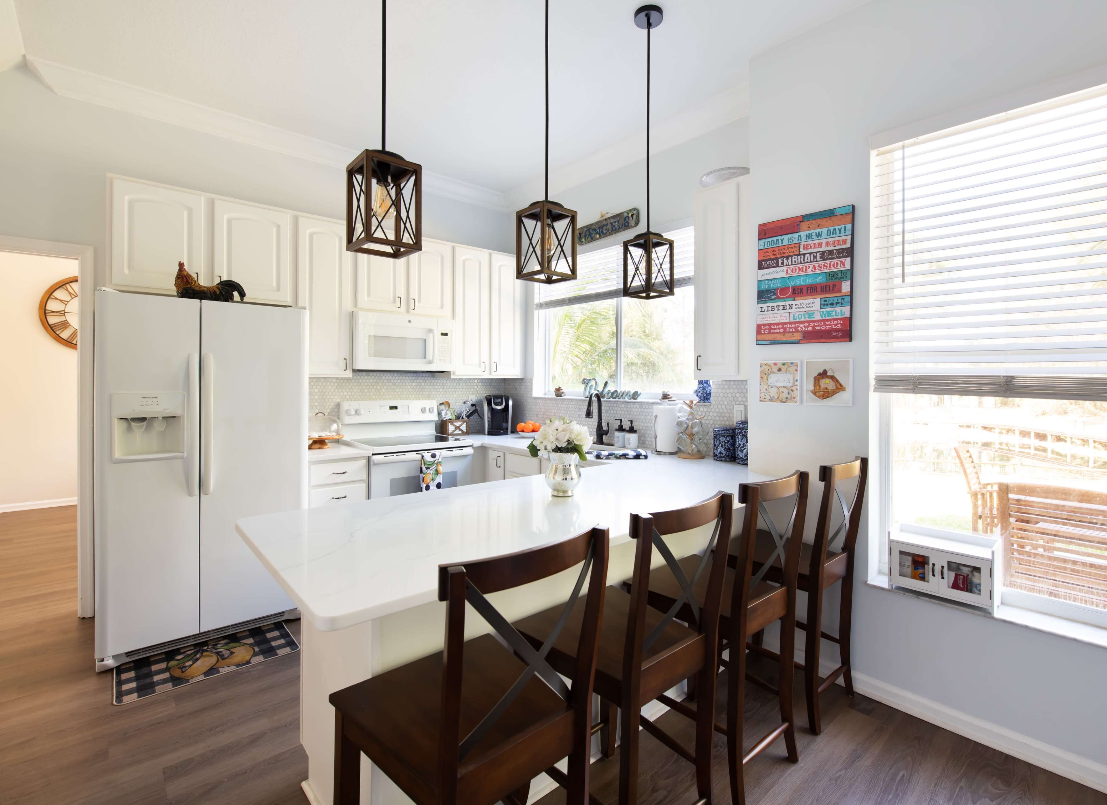 A modern kitchen features white cabinets, a central island with seating, and three pendant lights above, with large windows allowing natural light to enter.