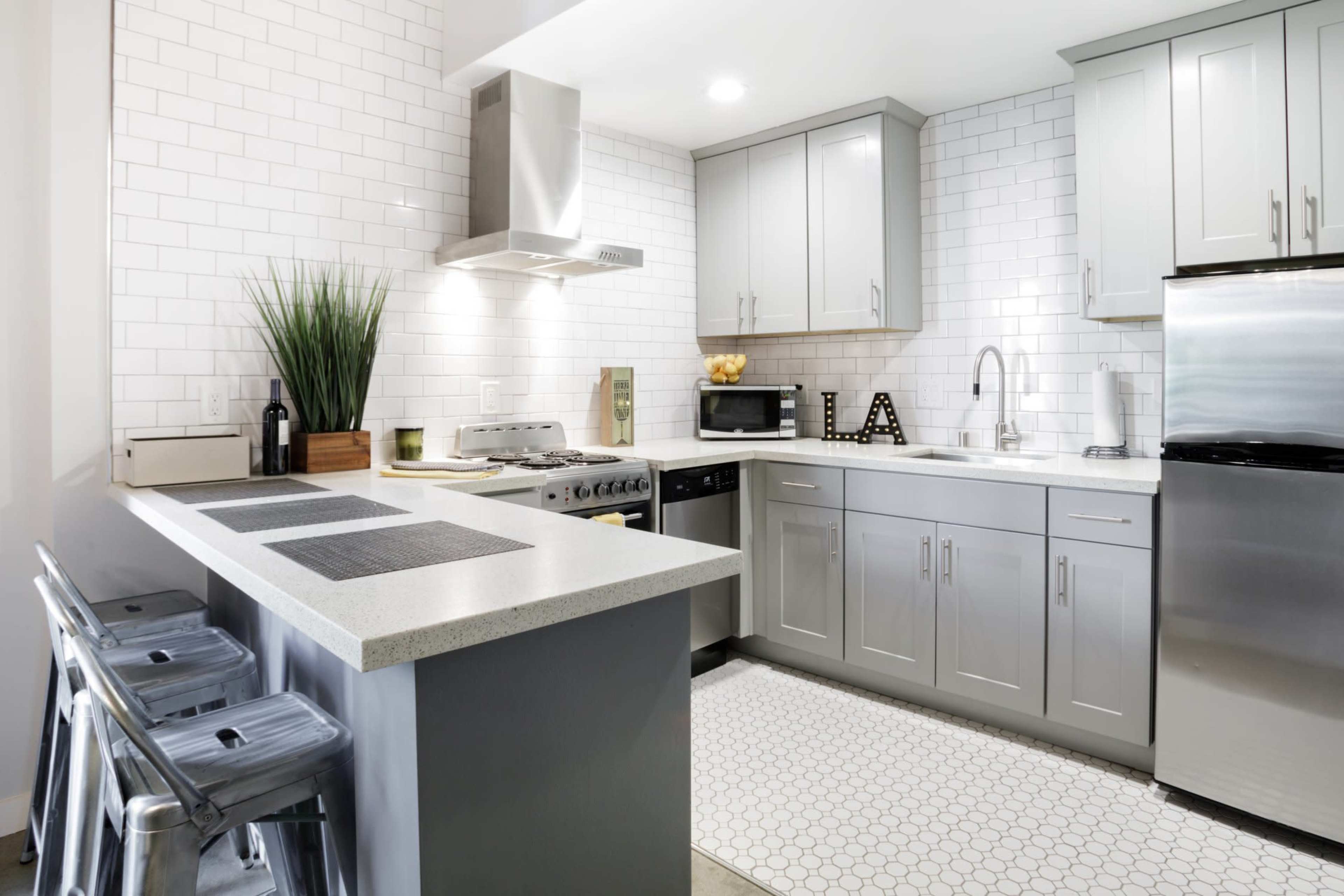 The image shows a modern kitchen with gray cabinets, a white tile backsplash, stainless steel appliances, and a hexagonal tiled floor.