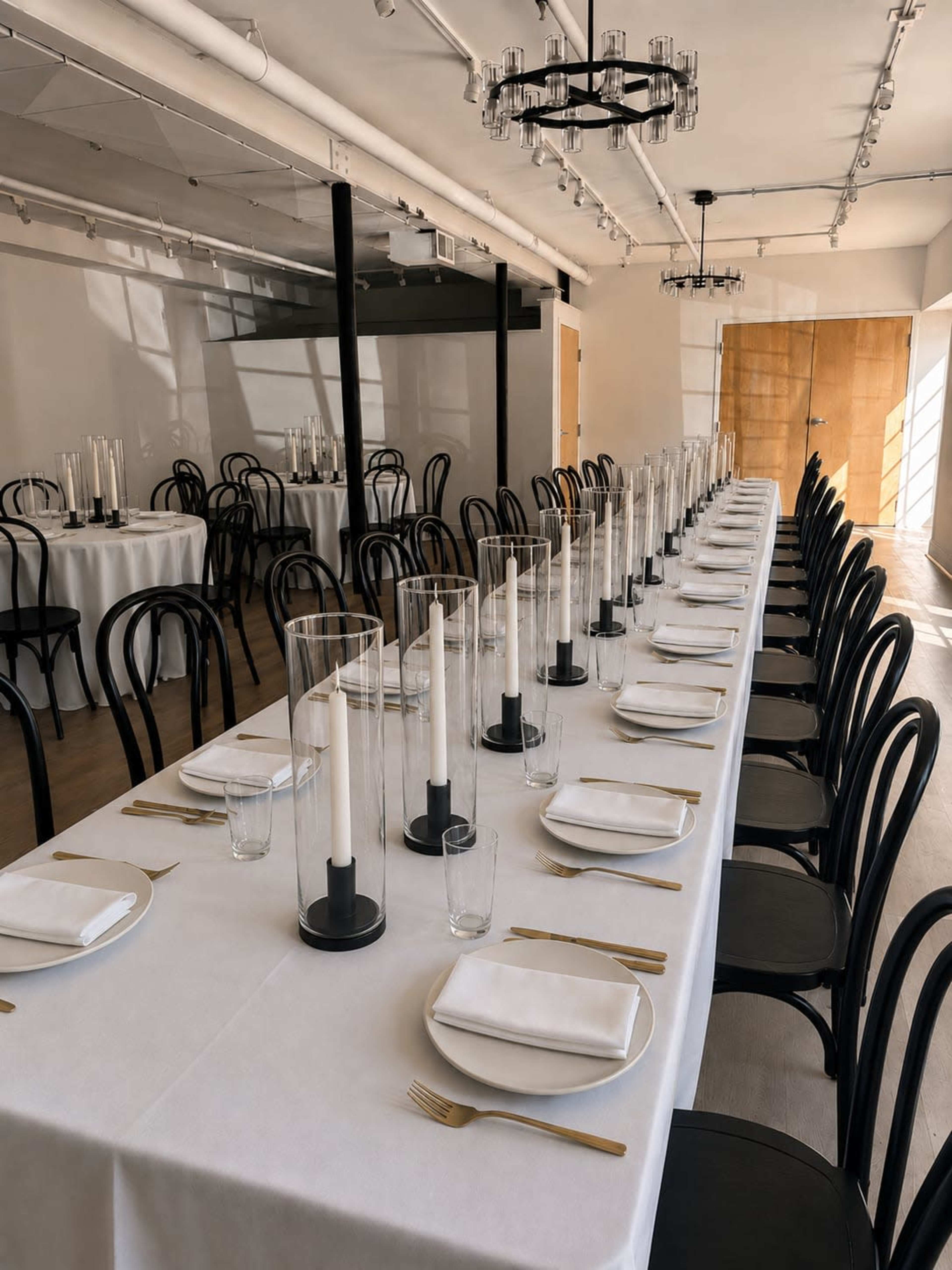 A long dining table is set with white tablecloths, clear glass candle holders, and gold utensils in a modern, well-lit space.