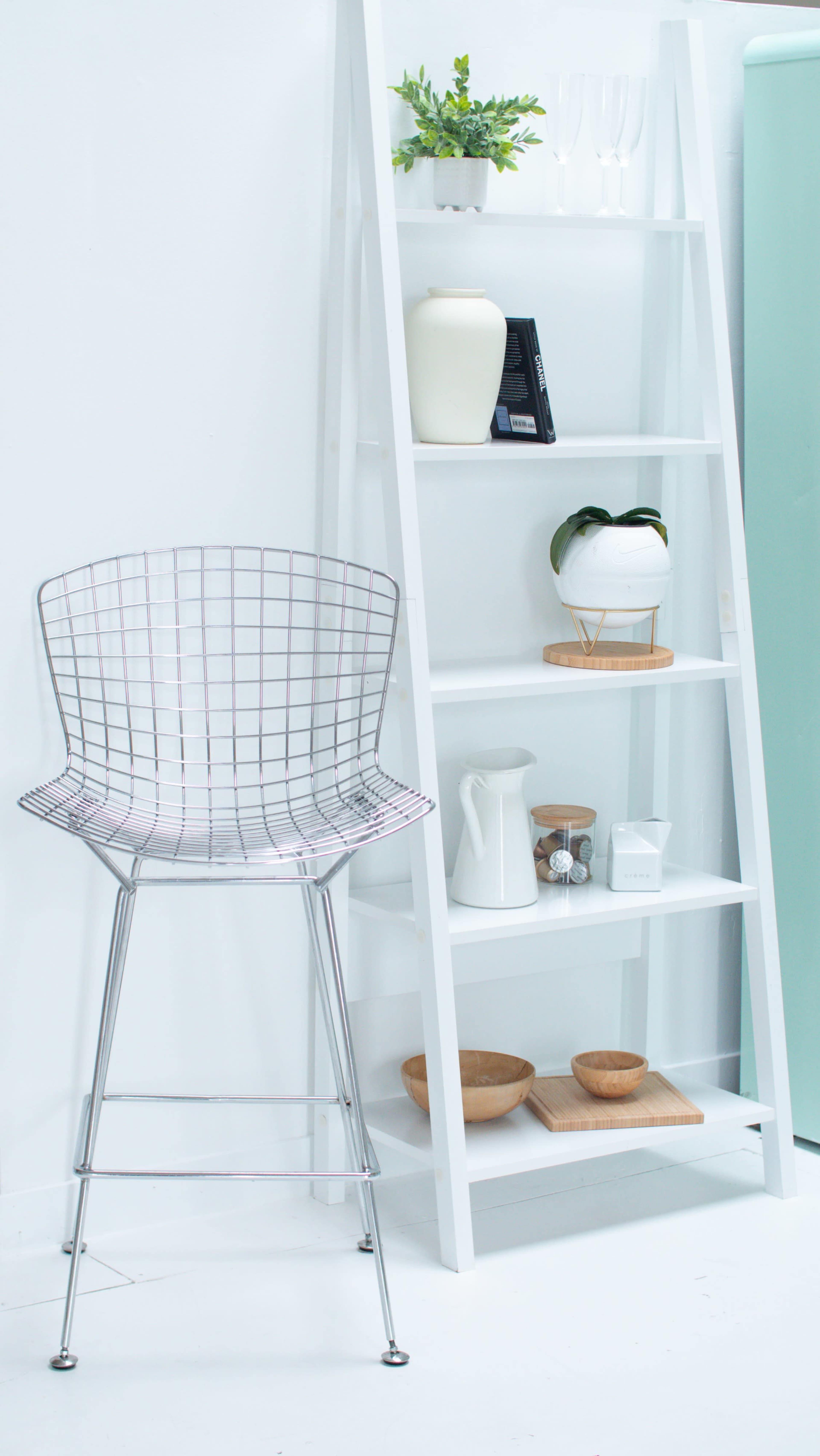 A wire bar stool stands next to a white shelving unit filled with various decorative items and plants.