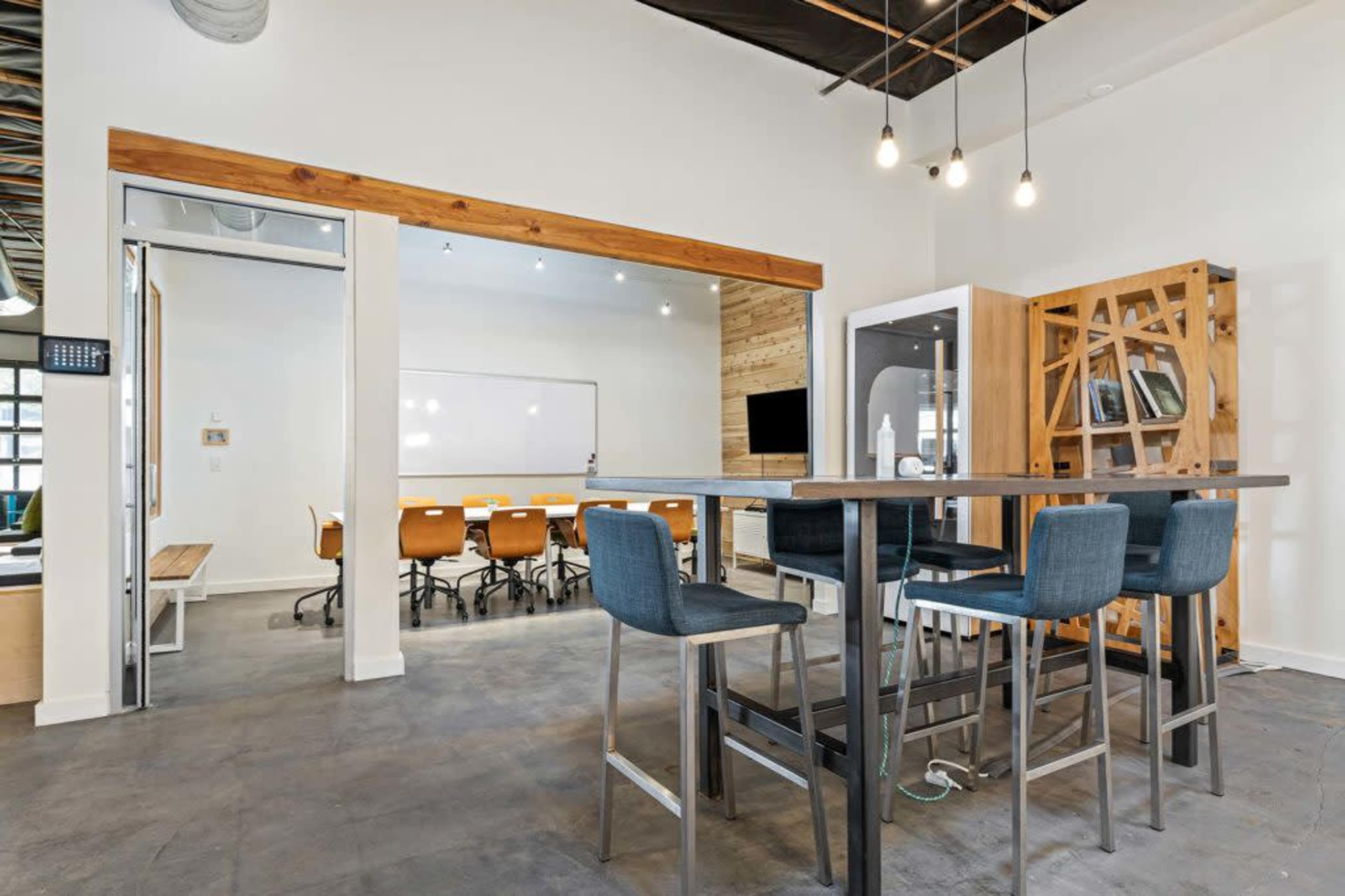 A modern office space features a high table with four chairs, a glass meeting room, and a large whiteboard in the background.