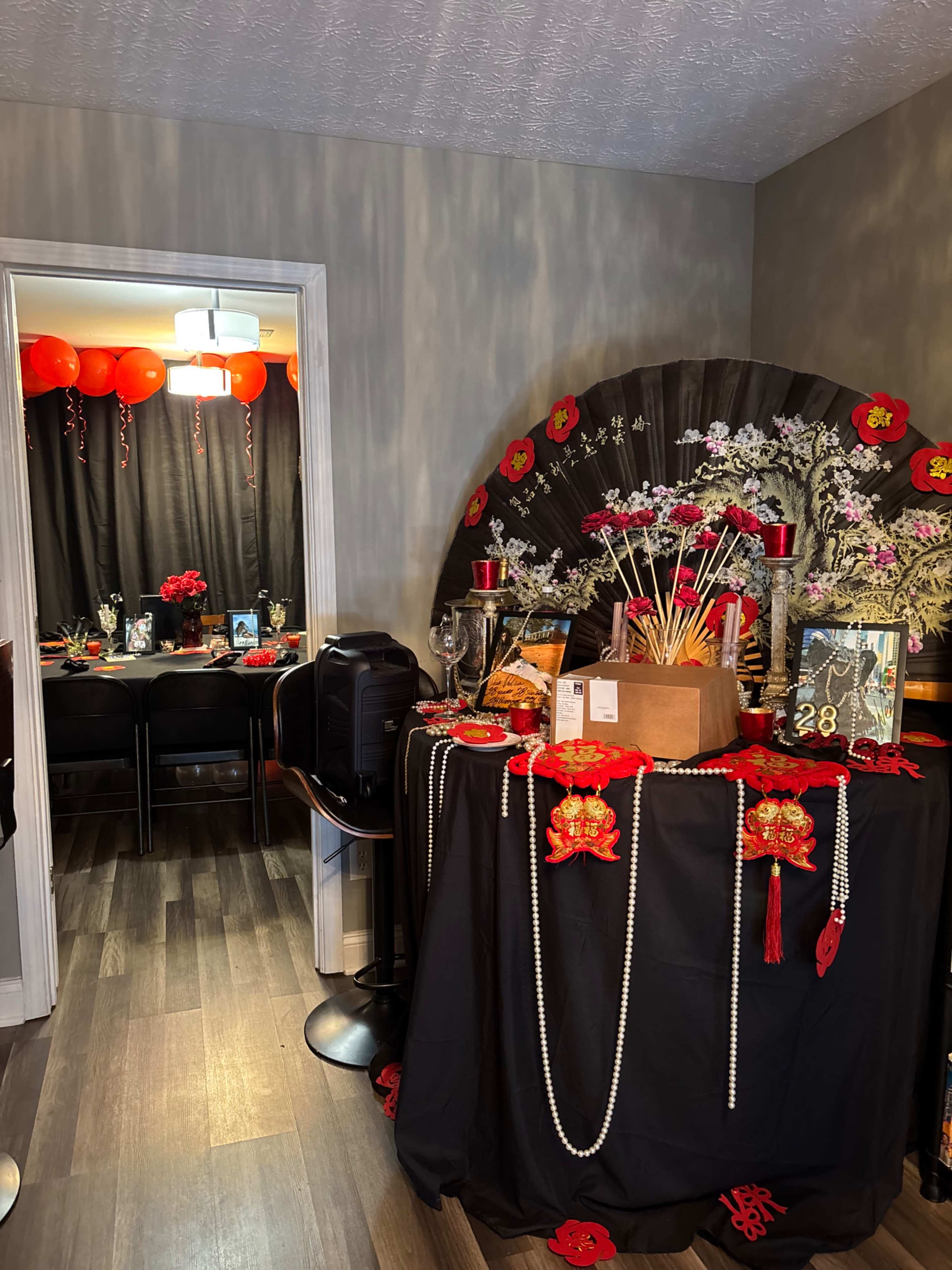 A decorated table with red and gold accents, flowers, and symbolic items is set up in a room leading to a festive gathering area adorned with lanterns and floral arrangements.