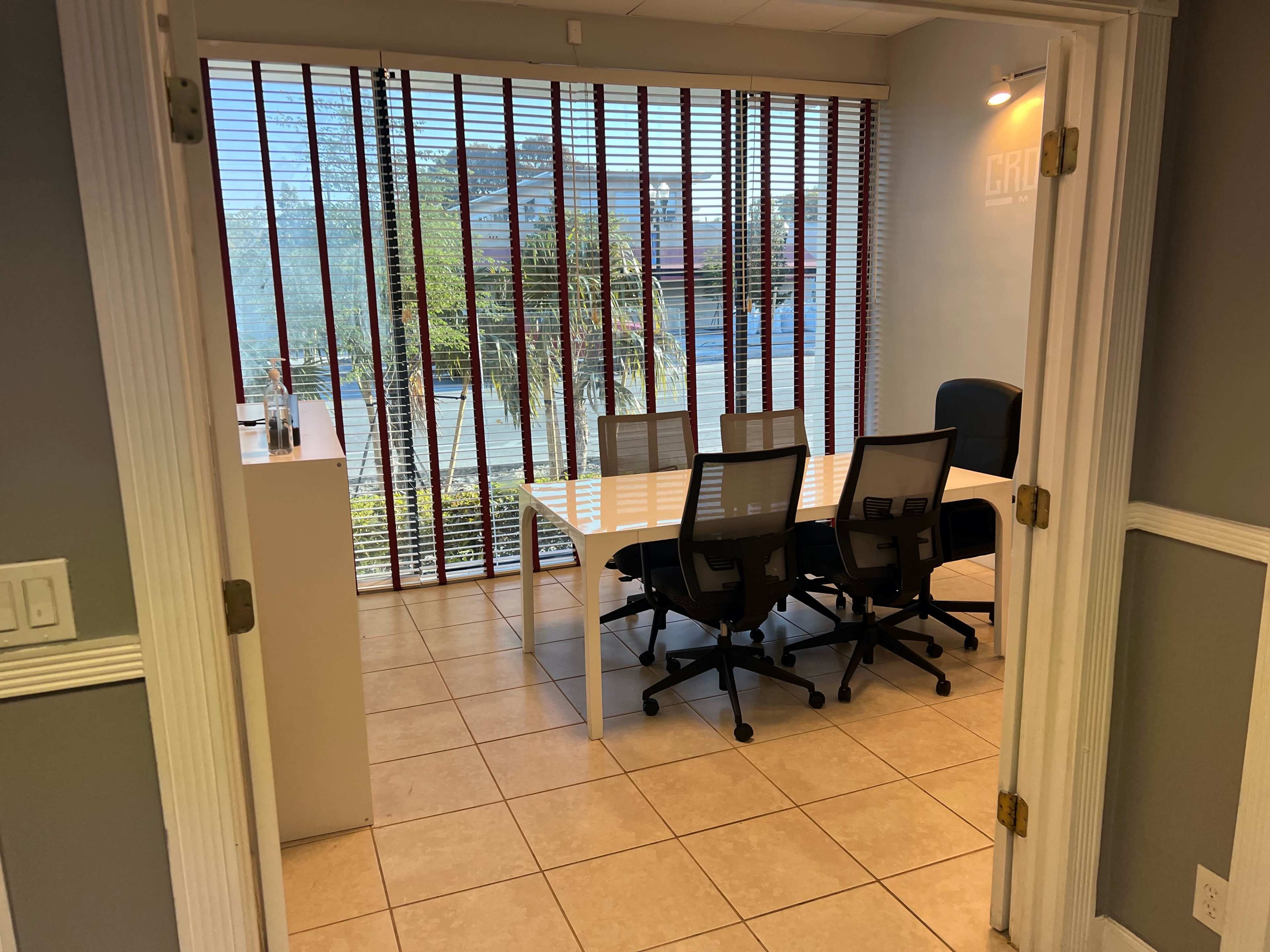The image shows a modern office space with a conference table and chairs, illuminated by natural light through vertical blinds.