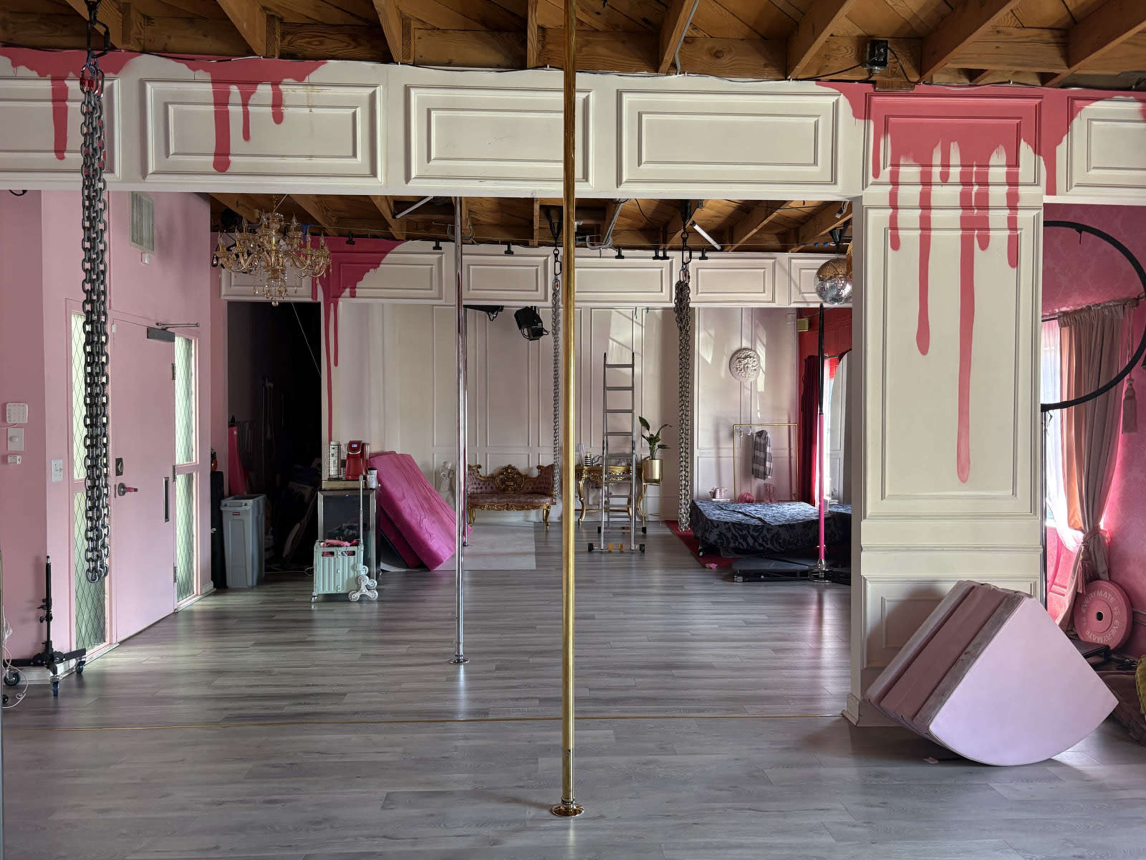 The image shows a spacious interior with a wooden ceiling, featuring a gold pole, pink accents on the walls, a bed in the background, and various furniture and equipment scattered throughout the room.