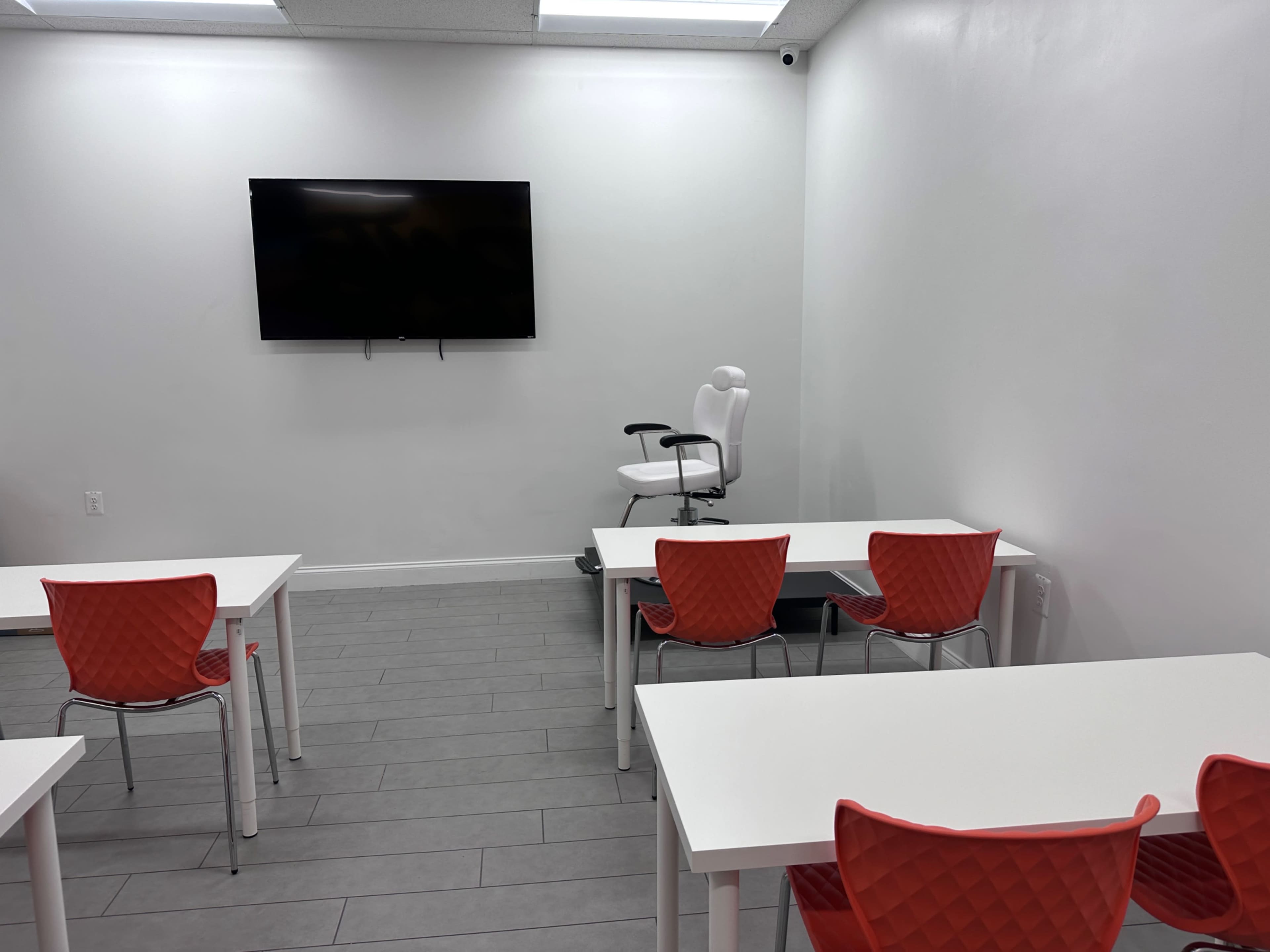 A modern classroom features white tables and red chairs, with a large wall-mounted screen and a single office chair positioned in the corner.