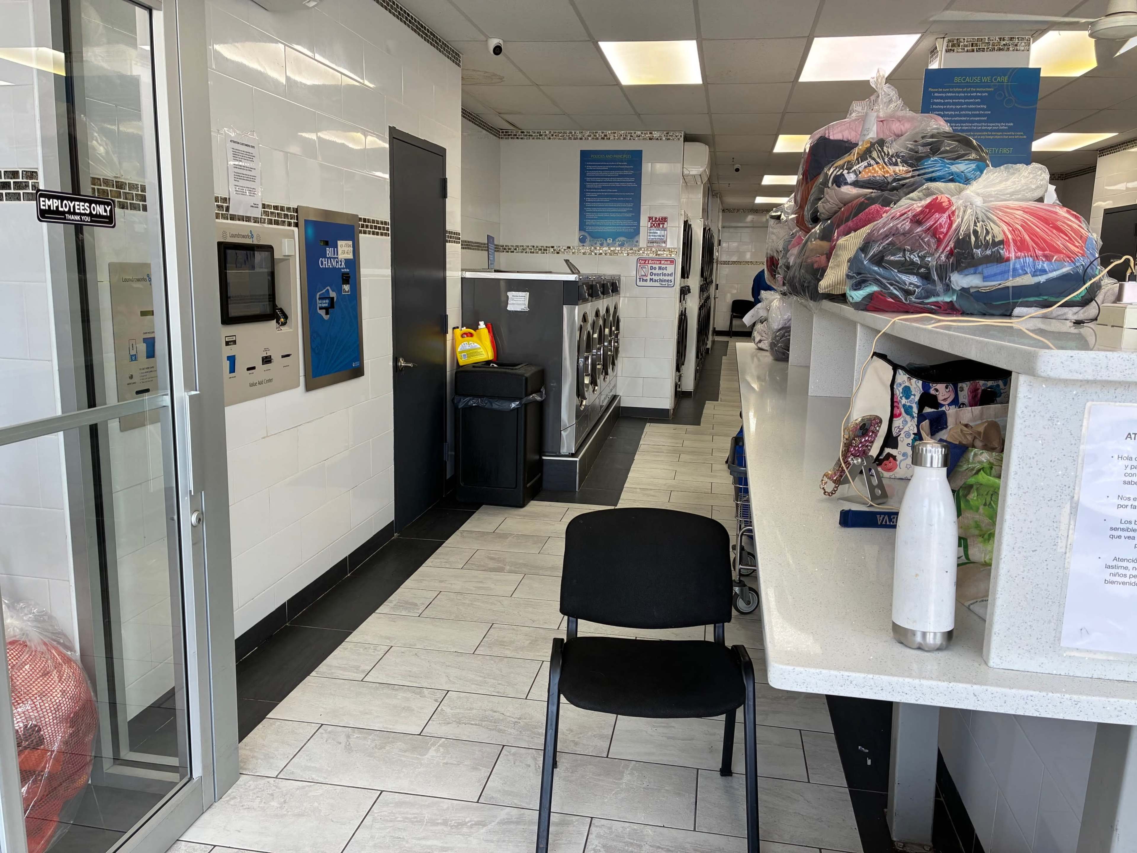 The image shows the interior of a laundromat with washing machines lining the walls, a chair in the foreground, and a pile of laundry on a counter.