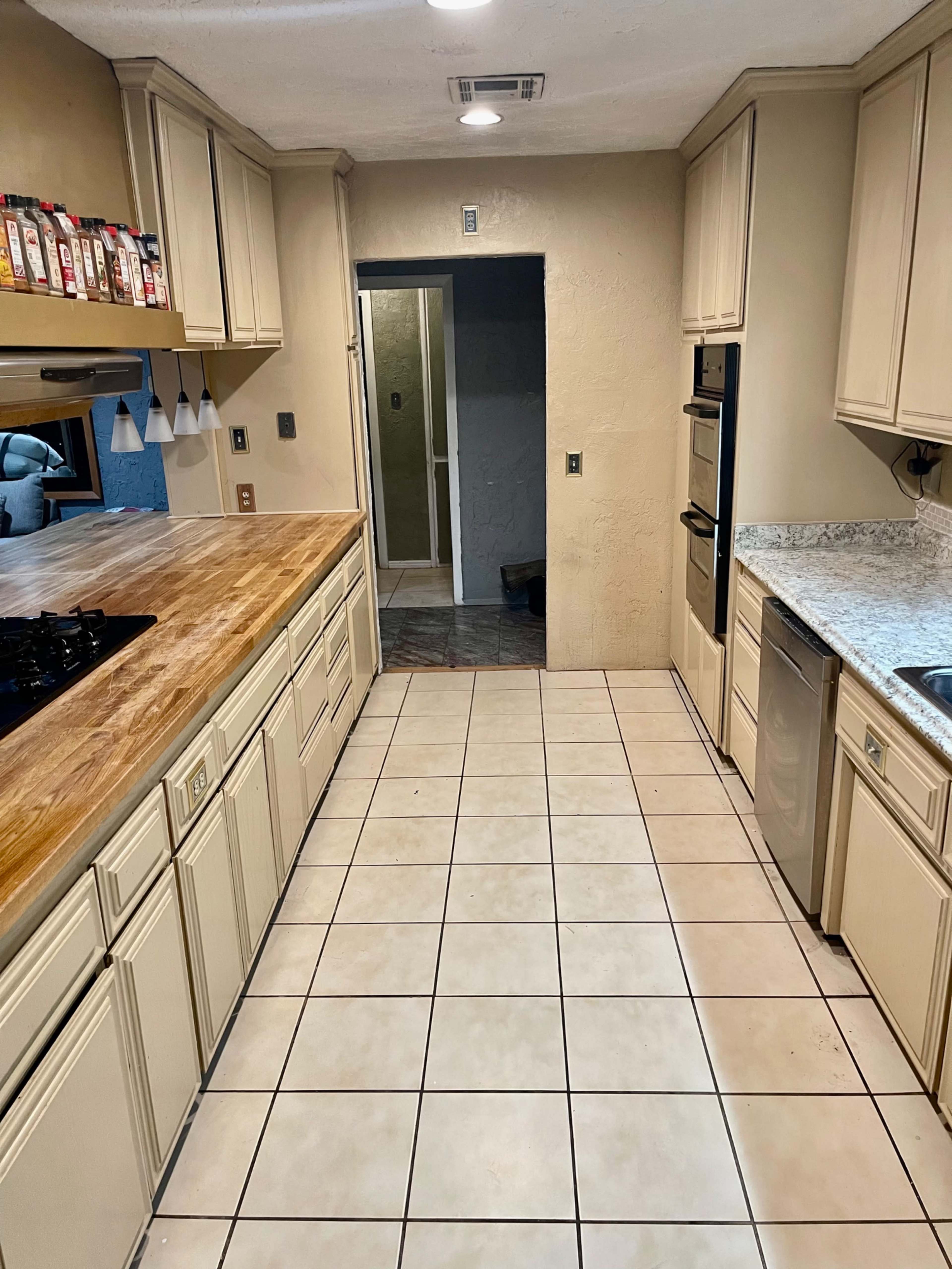 A kitchen with beige cabinets, a wooden countertop, stainless steel appliances, and tiled flooring, leading to an entryway with a bathroom visible in the background.
