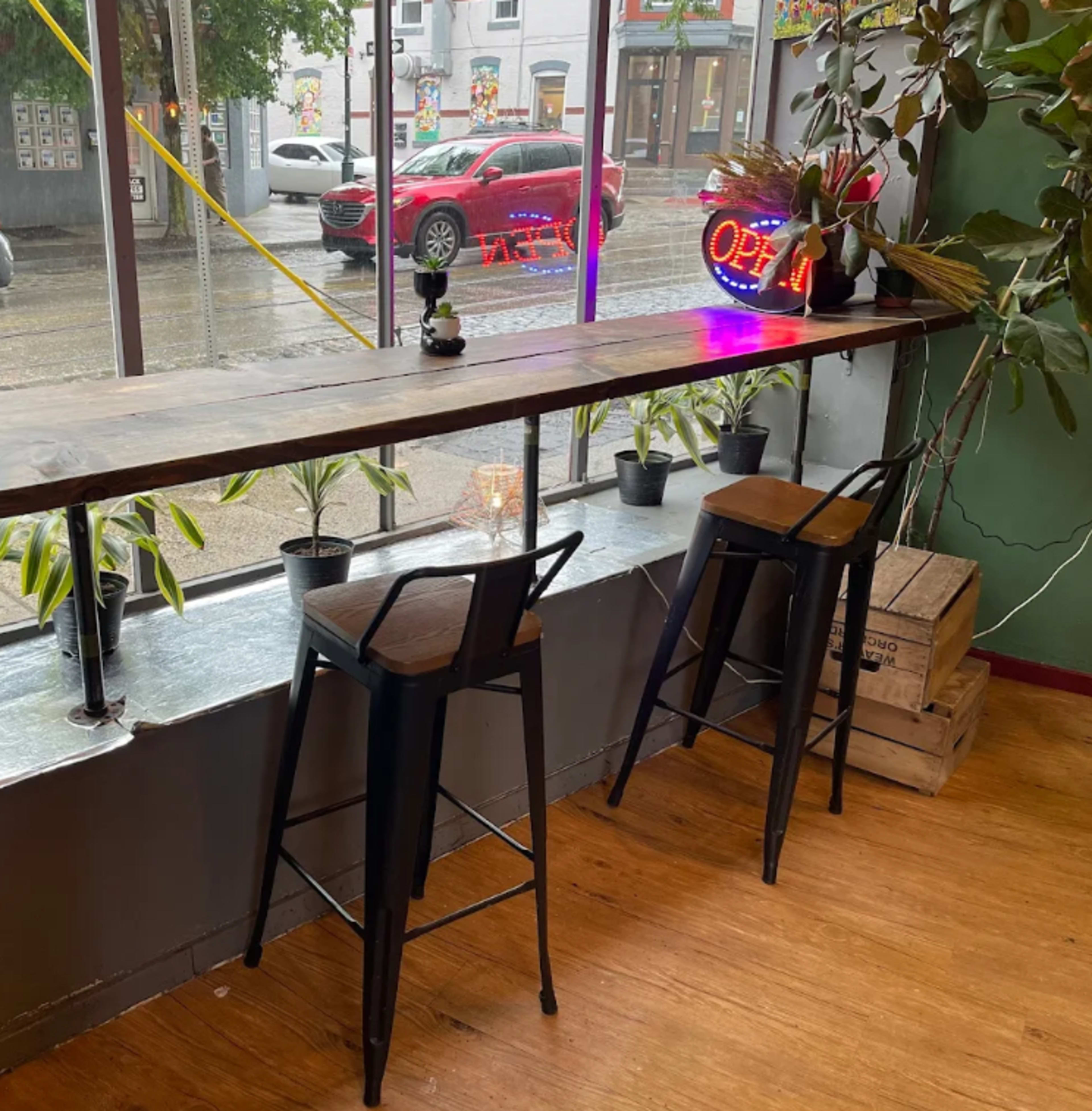 The image shows a modern cafe interior with a wooden bar countertop, two black bar stools, and potted plants near large windows.