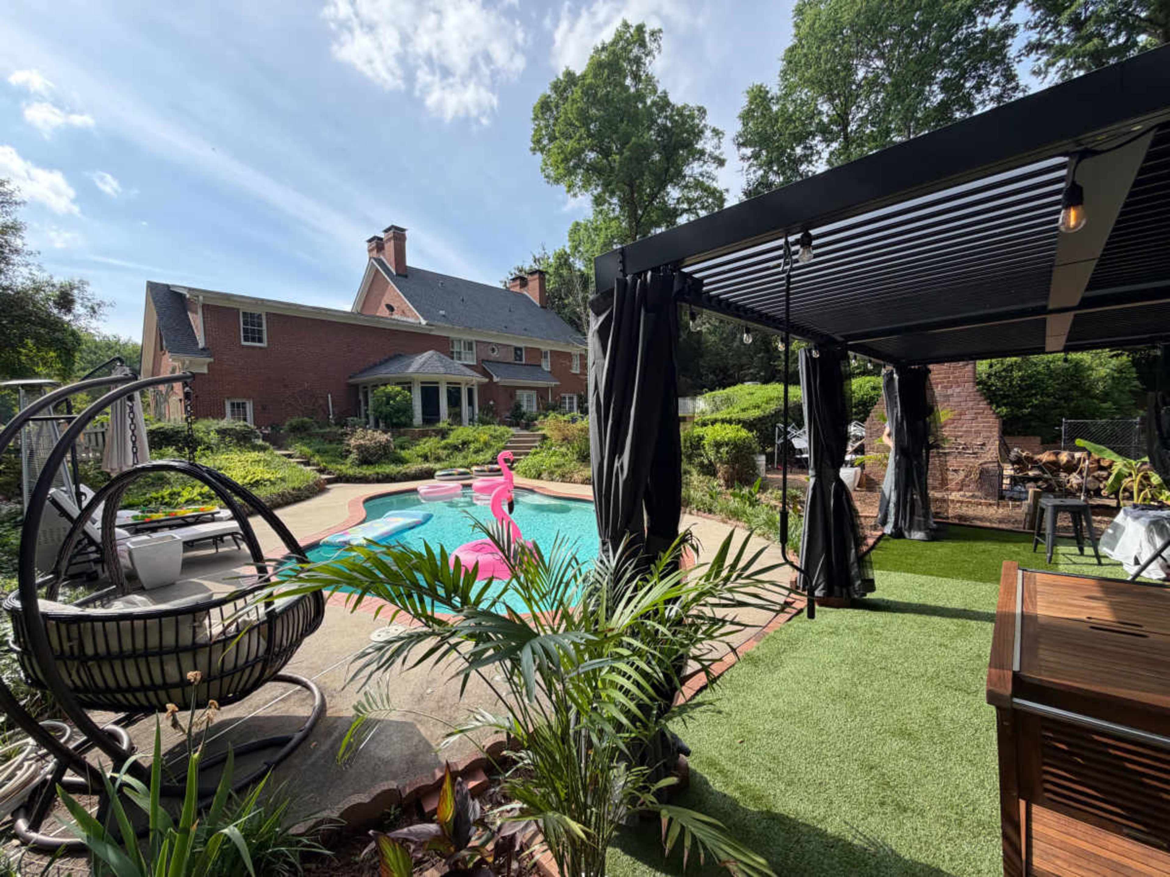 The image shows a backyard with a swimming pool, a pink flamingo float, landscaped greenery, and a covered seating area adjacent to a large brick house.