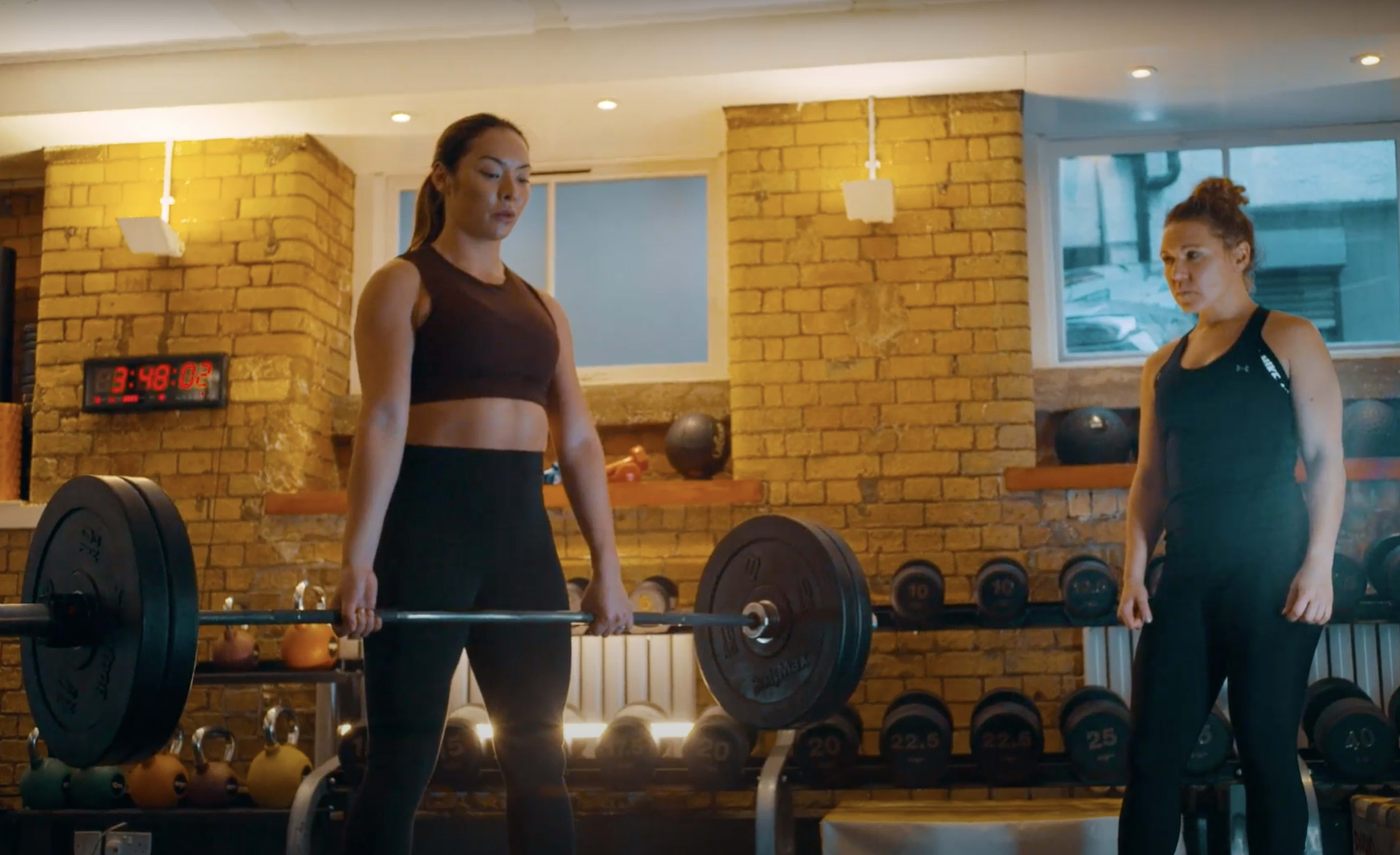Two women are in a gym setting, with one lifting a barbell while the other observes from the side.