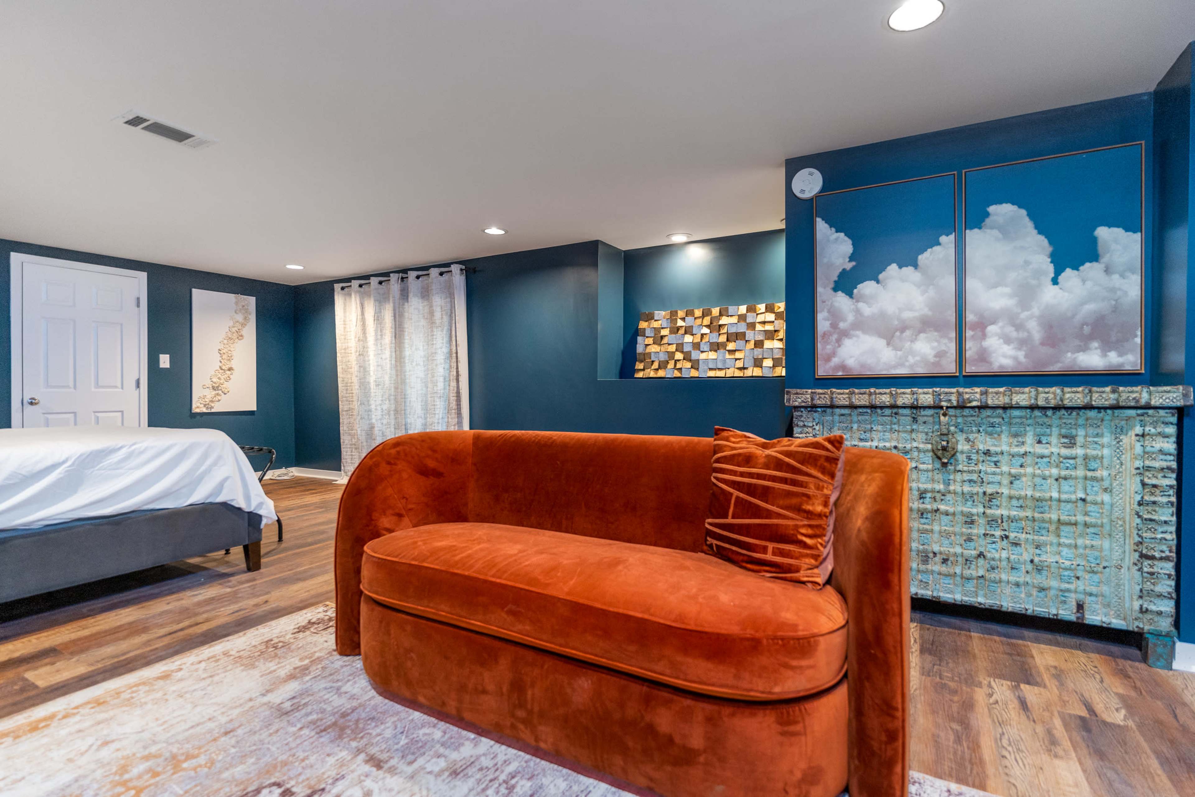 A modern room featuring a curved orange sofa, a bed with white bedding, decorative wall art, and an accent wall colored in dark blue.