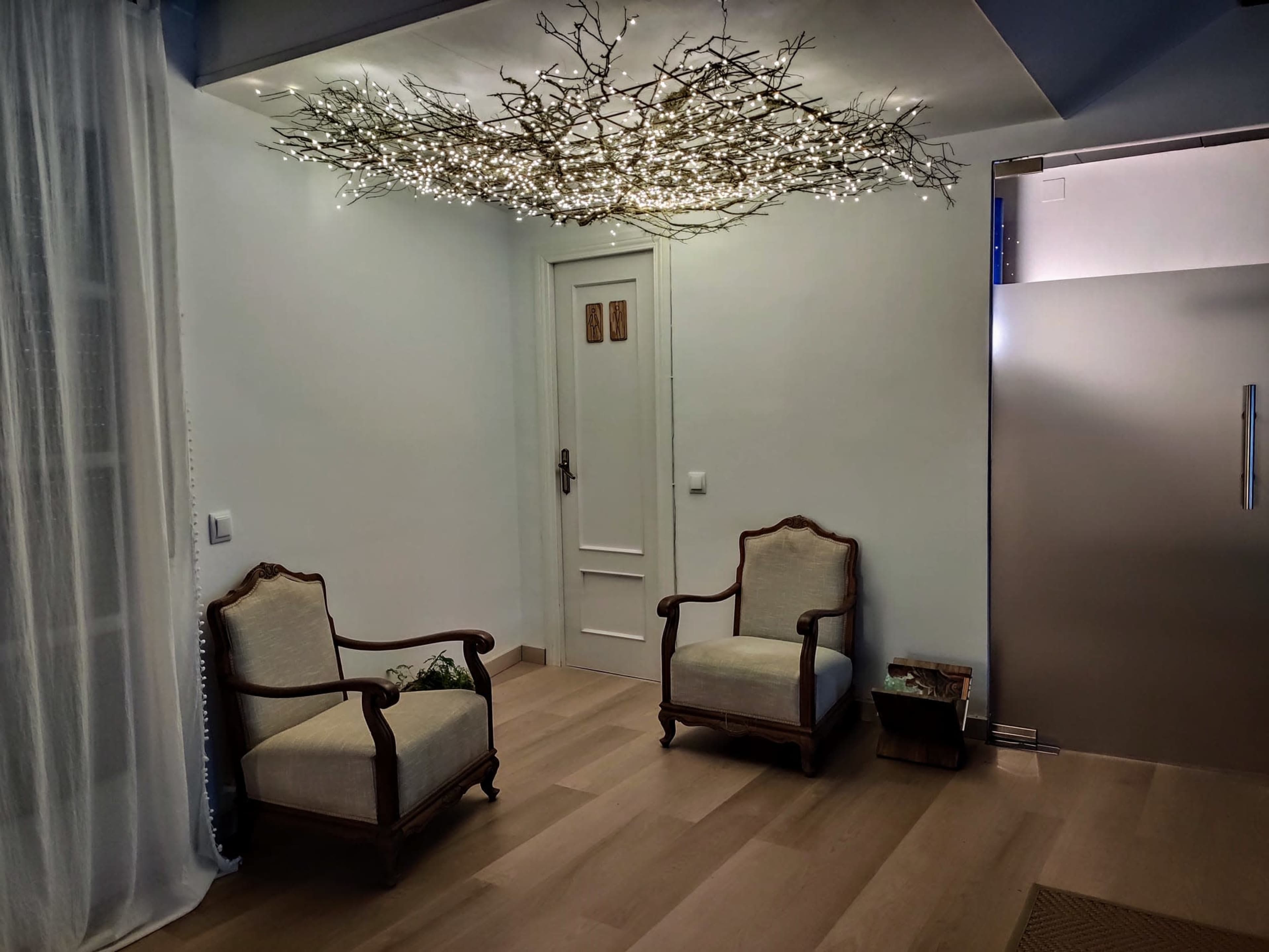 A room with two wooden chairs facing each other, illuminated by a decorative light fixture resembling branches above, and a fridge visible in the background.