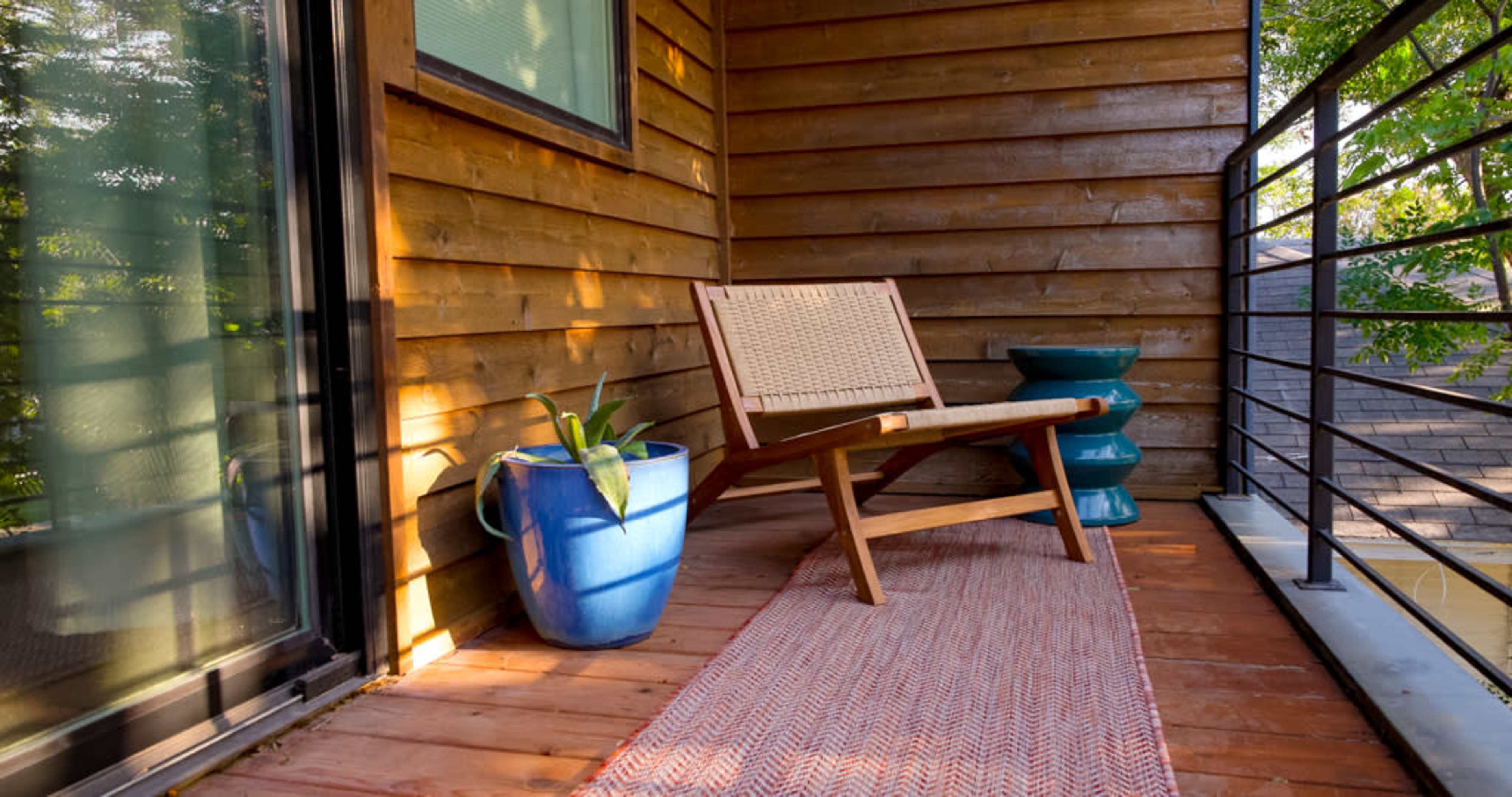 A wooden balcony features a chair, a blue flower pot with a plant, a small blue table, and a woven rug.