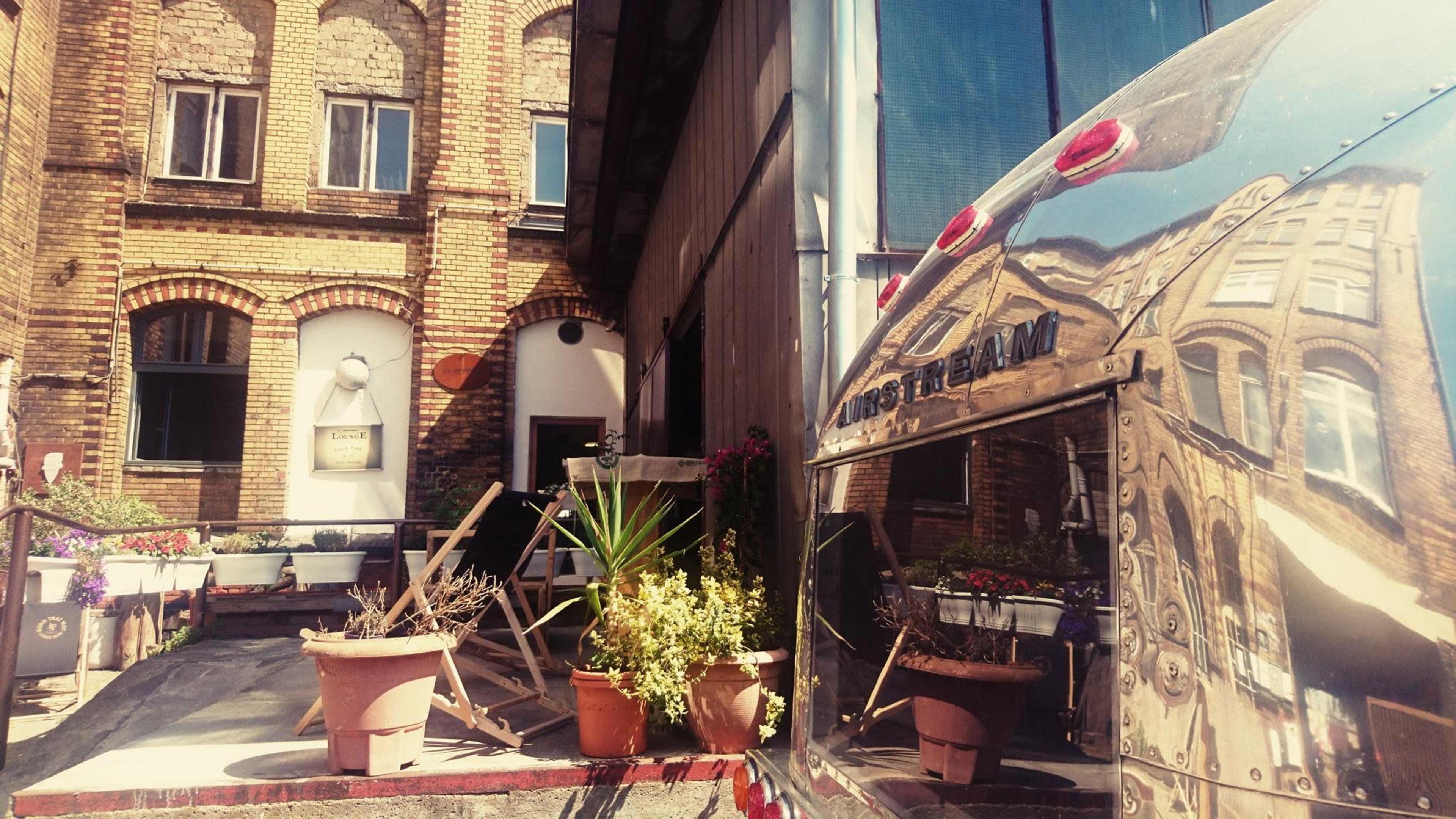 A brick building entrance with potted plants and an Airstream trailer reflected in the foreground.
