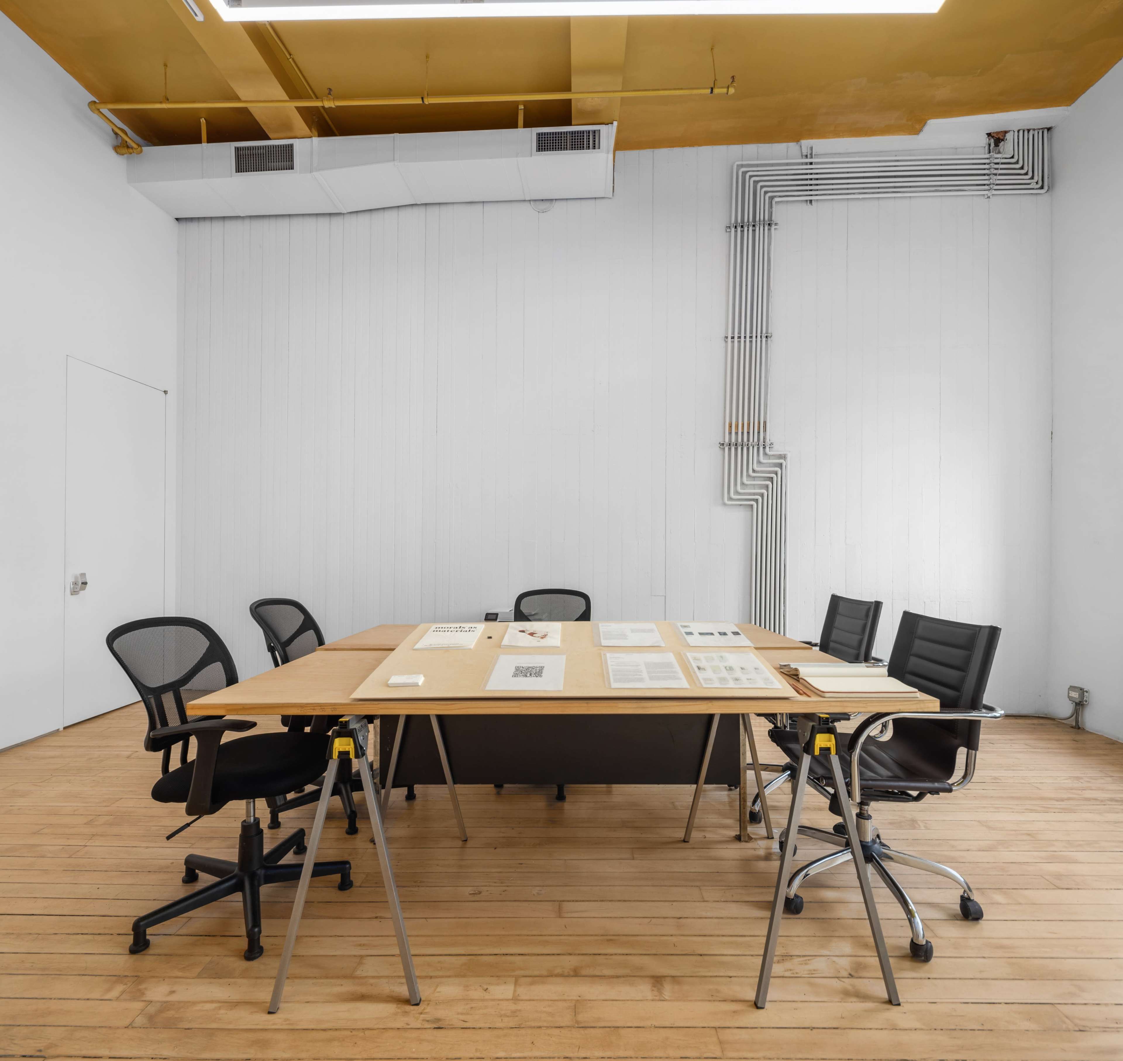 A conference room features a large wooden table surrounded by black office chairs and a plain white wall with visible electrical conduits.