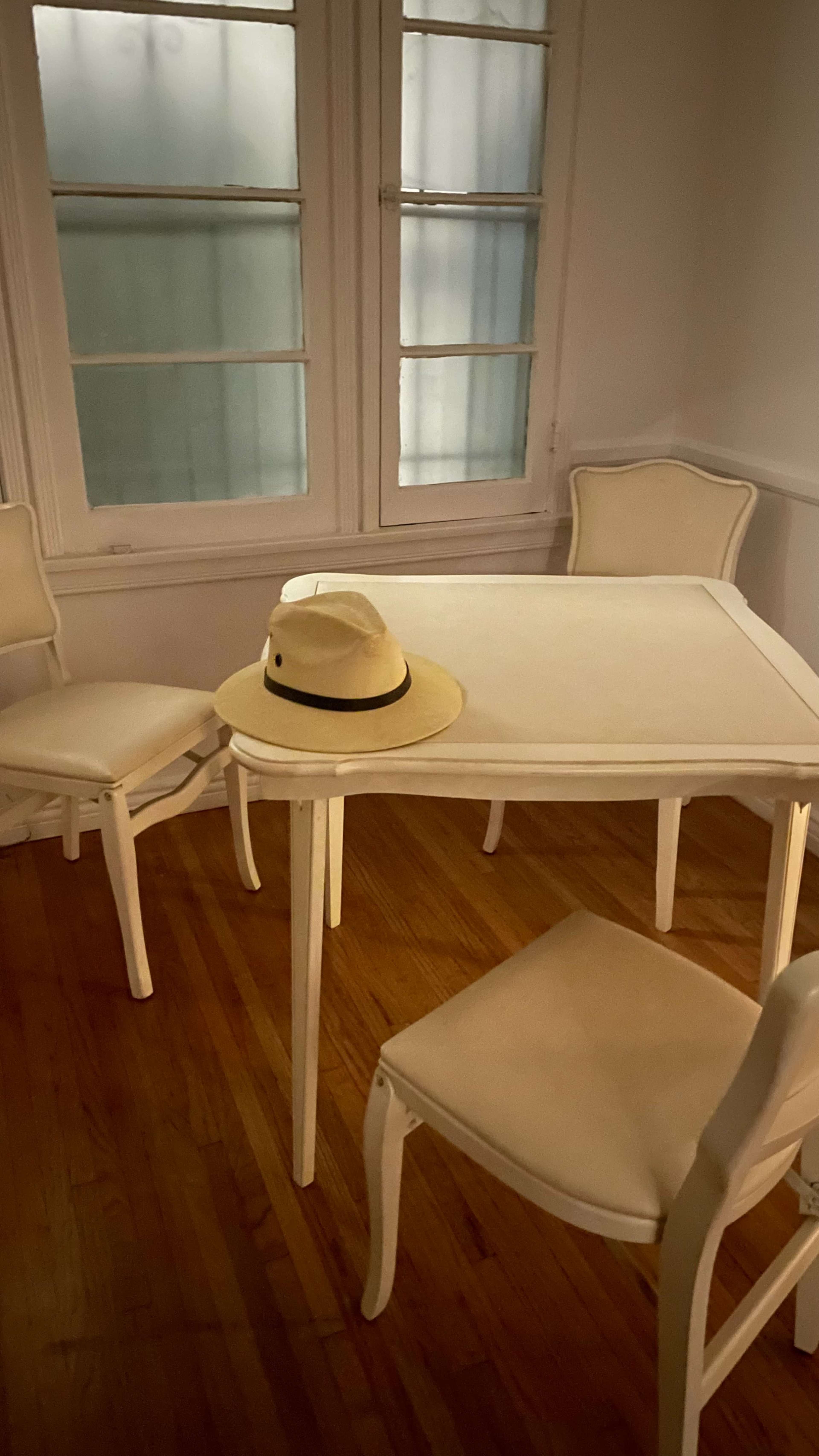 A white table with four matching chairs sits in a well-lit room, featuring a straw hat placed on the table.