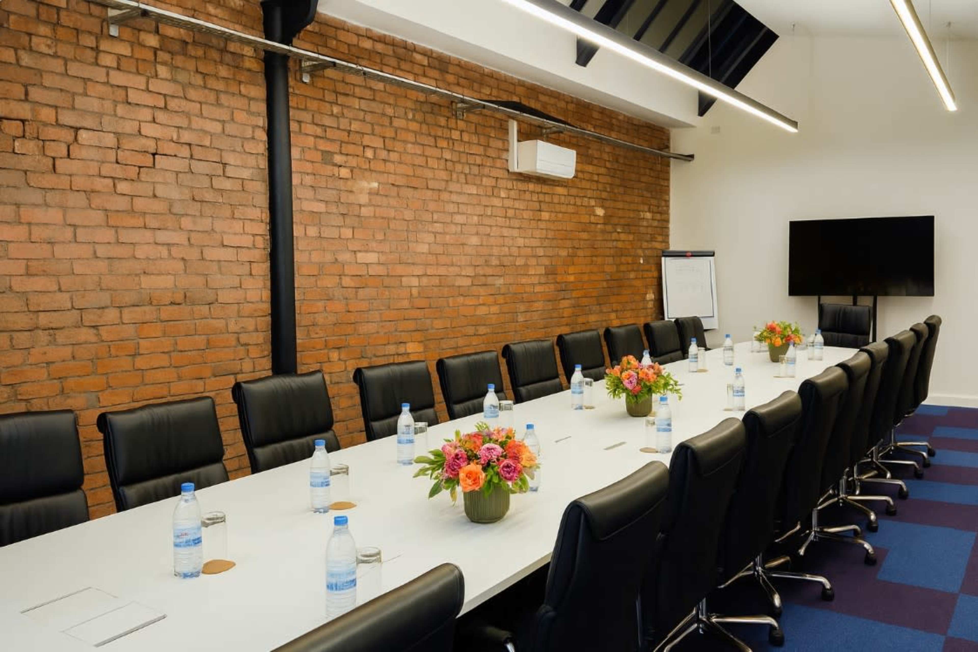 A large conference room features a long table with black chairs, decorated with floral centerpieces and water bottles, against a backdrop of exposed brick walls.