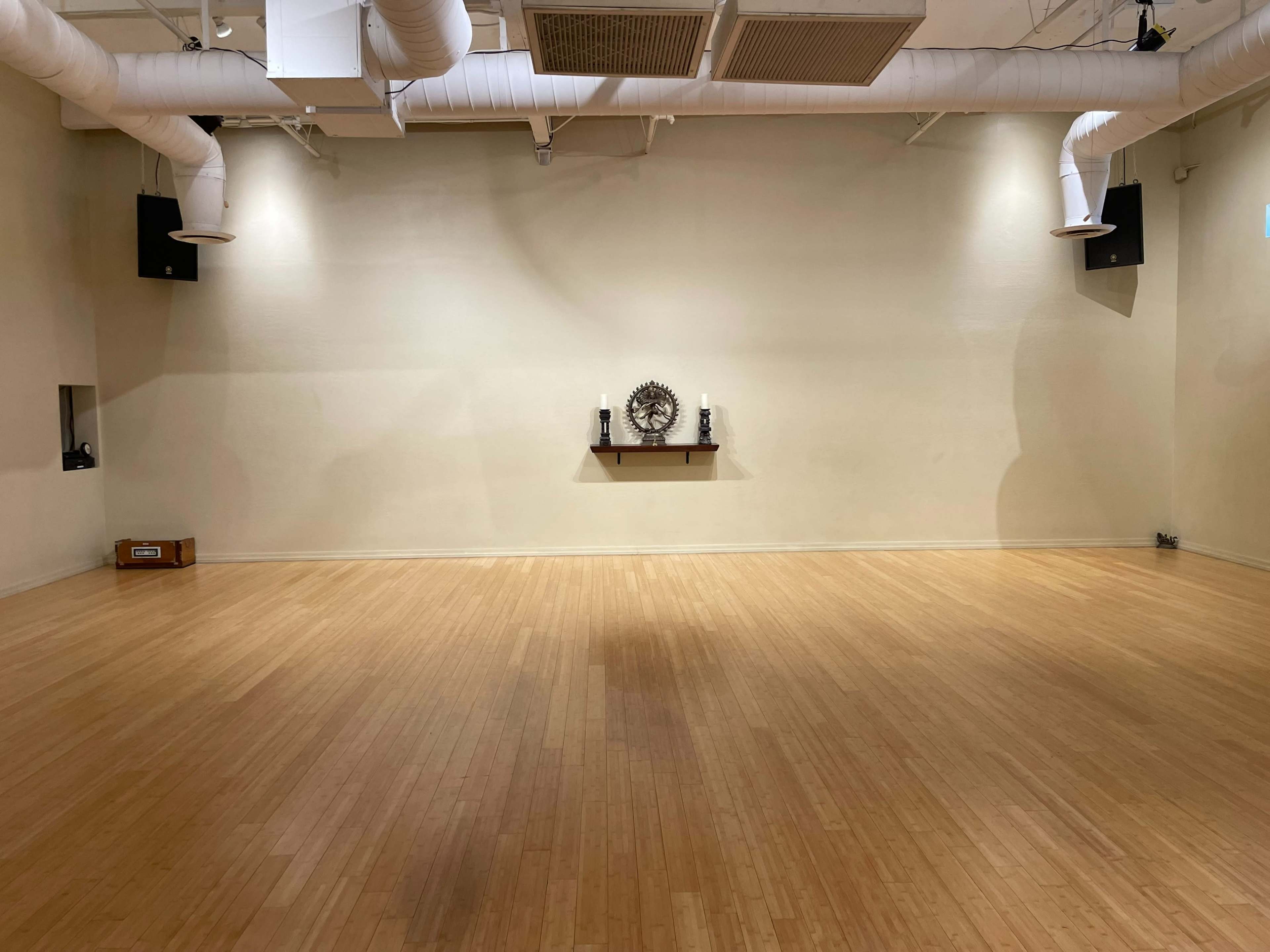 An empty wooden-floored studio with a plain wall, a shelf with decorative items in the center, and speakers mounted in the corners.