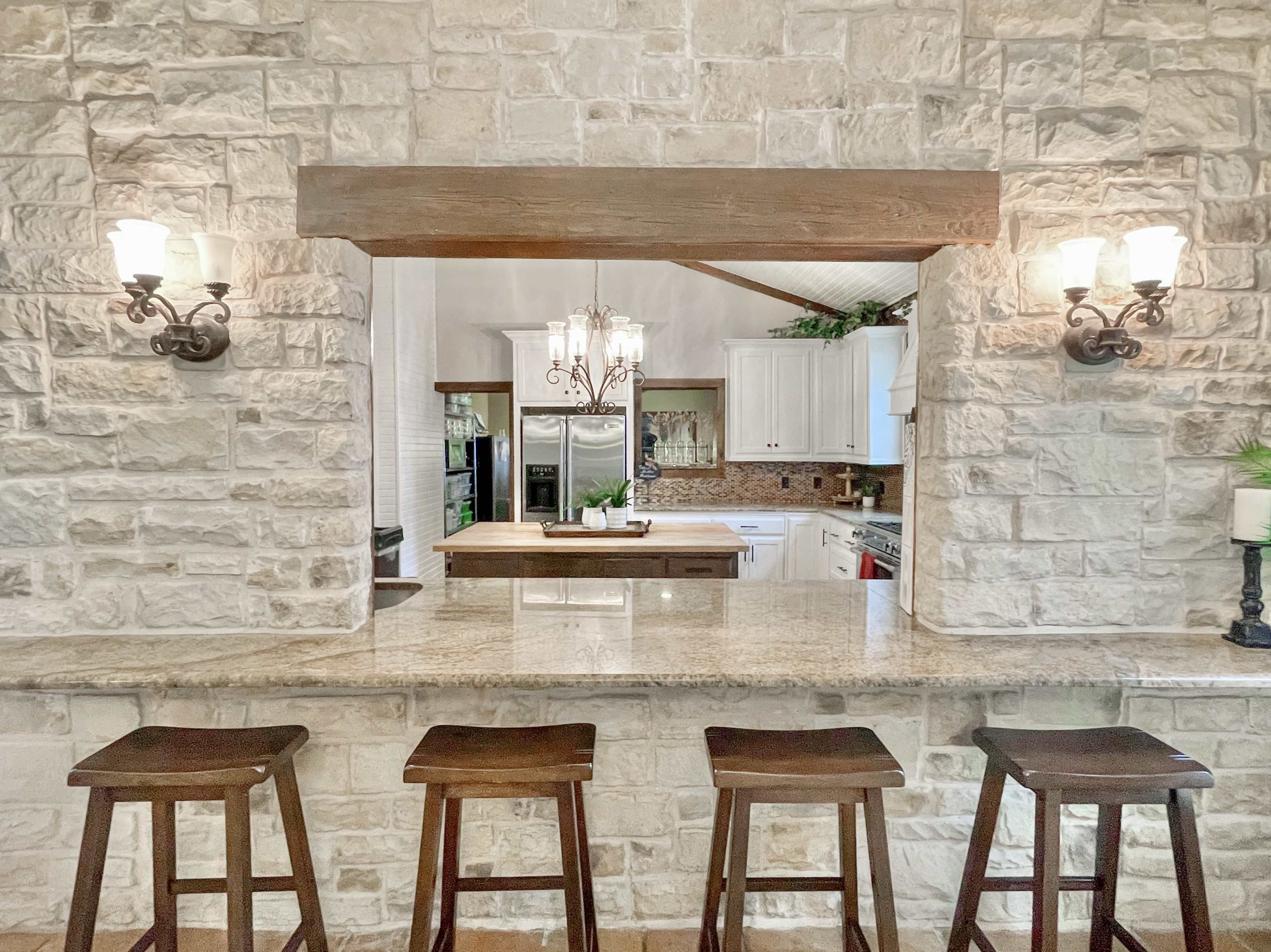 A stone-walled bar area with four wooden stools overlooks a kitchen featuring white cabinetry and a large island.