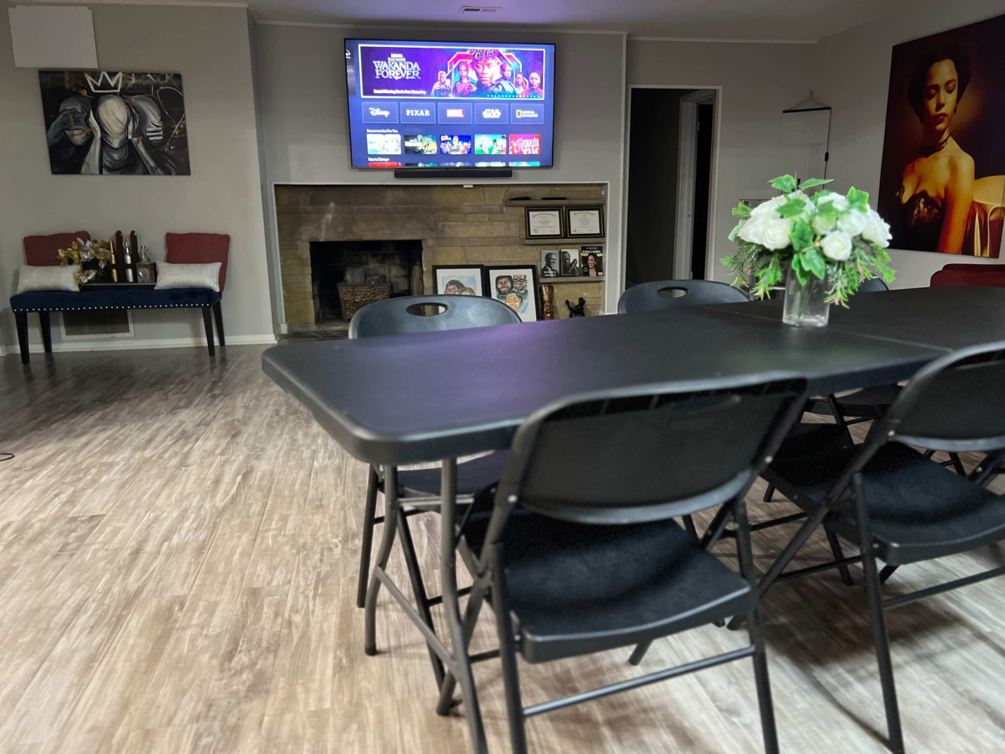 The image shows a spacious room with a black folding table and chairs, a stone fireplace, and a large television displaying various streaming options on the wall.