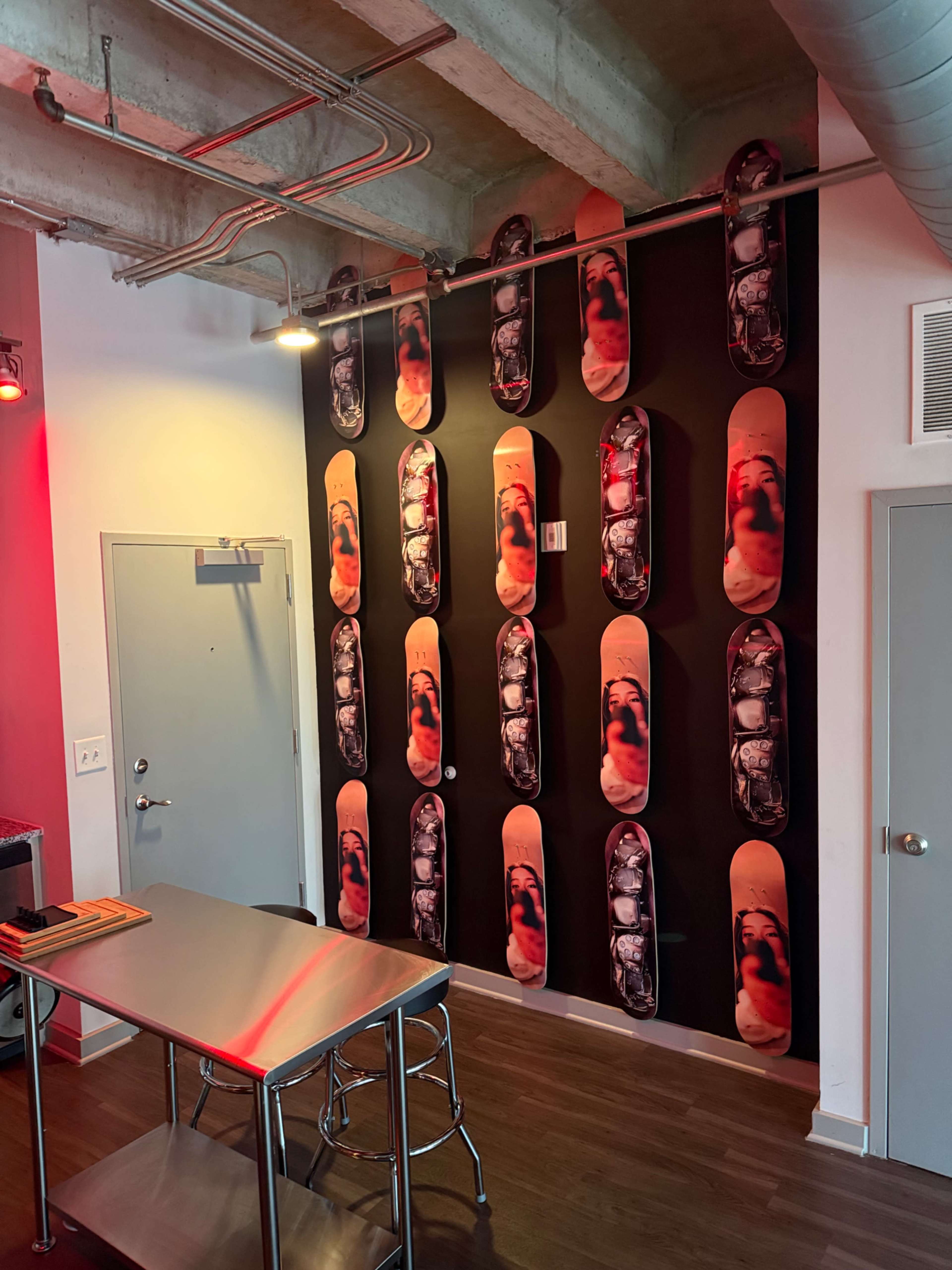 The image shows a modern interior with a wall decorated with skateboard decks arranged in a grid pattern, and a metal table and stools in the foreground.